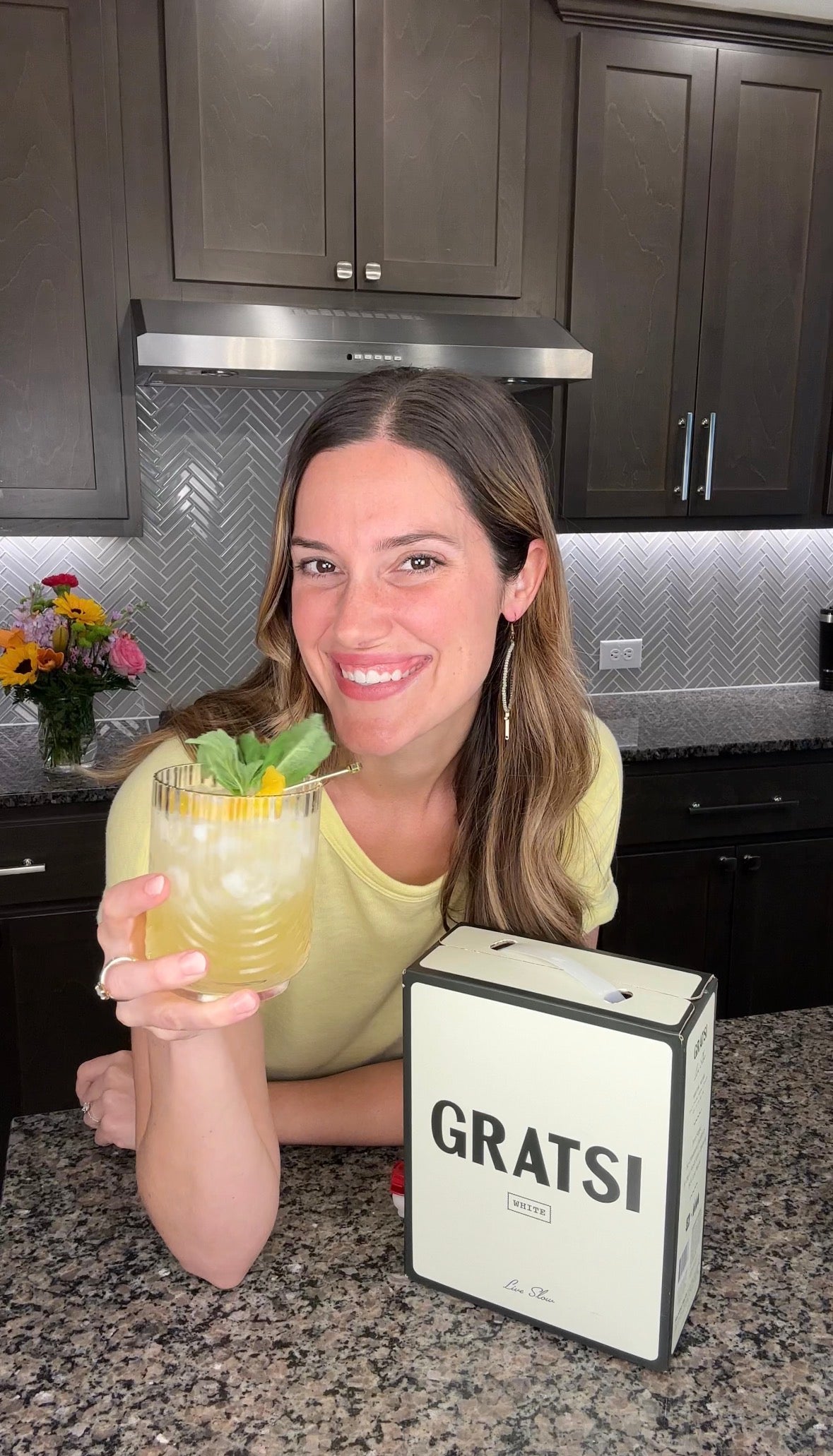 A woman with long brown hair smiles while holding a drink with mint and lemon in a glass. She stands in a modern kitchen next to a cream-colored box labeled GRATSI. Flowers are on the counter behind her.