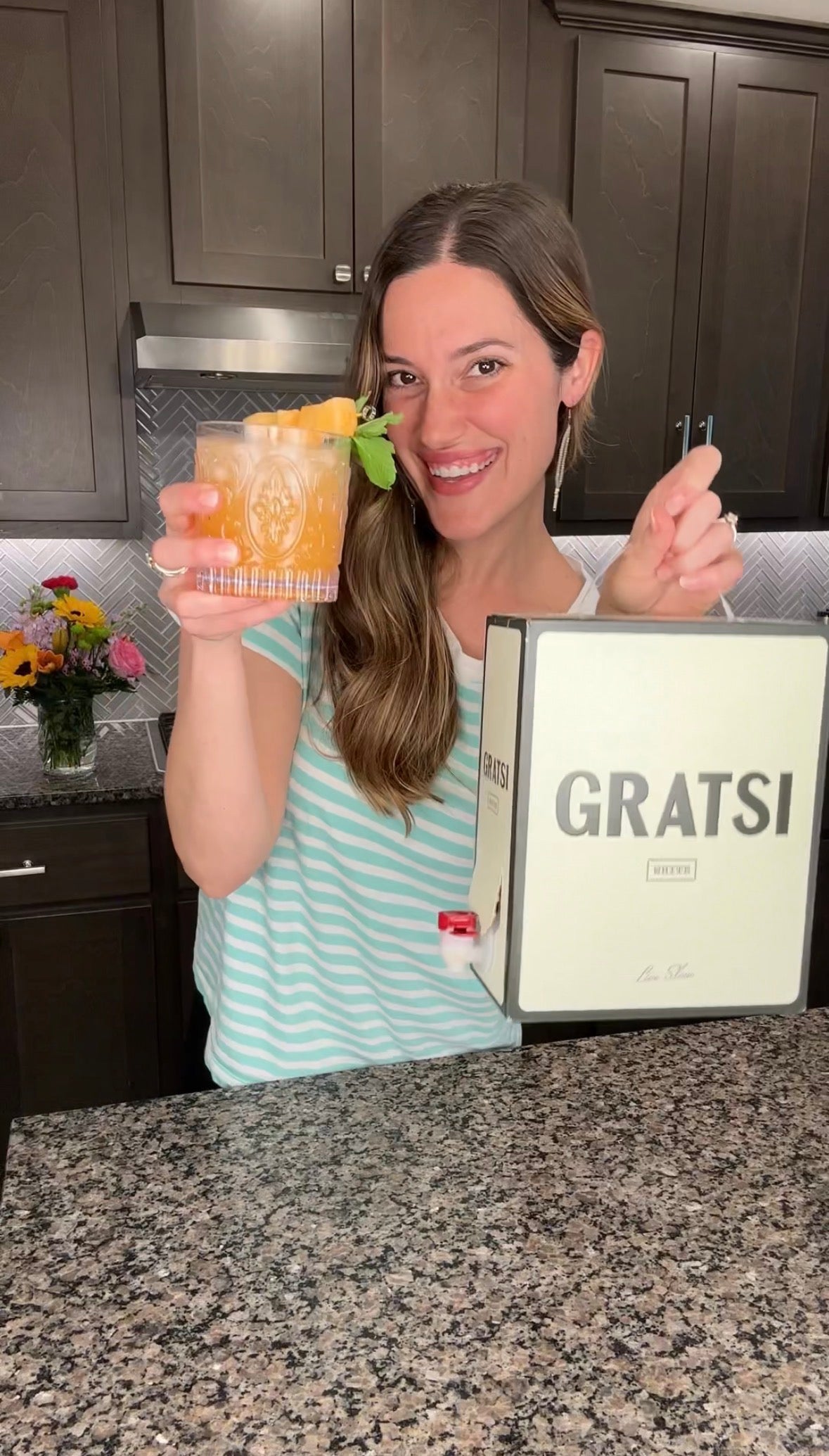 A woman in a striped shirt stands in a kitchen, smiling and holding up a cocktail and a box labeled GRATSI. A vase of flowers is on the counter behind her.