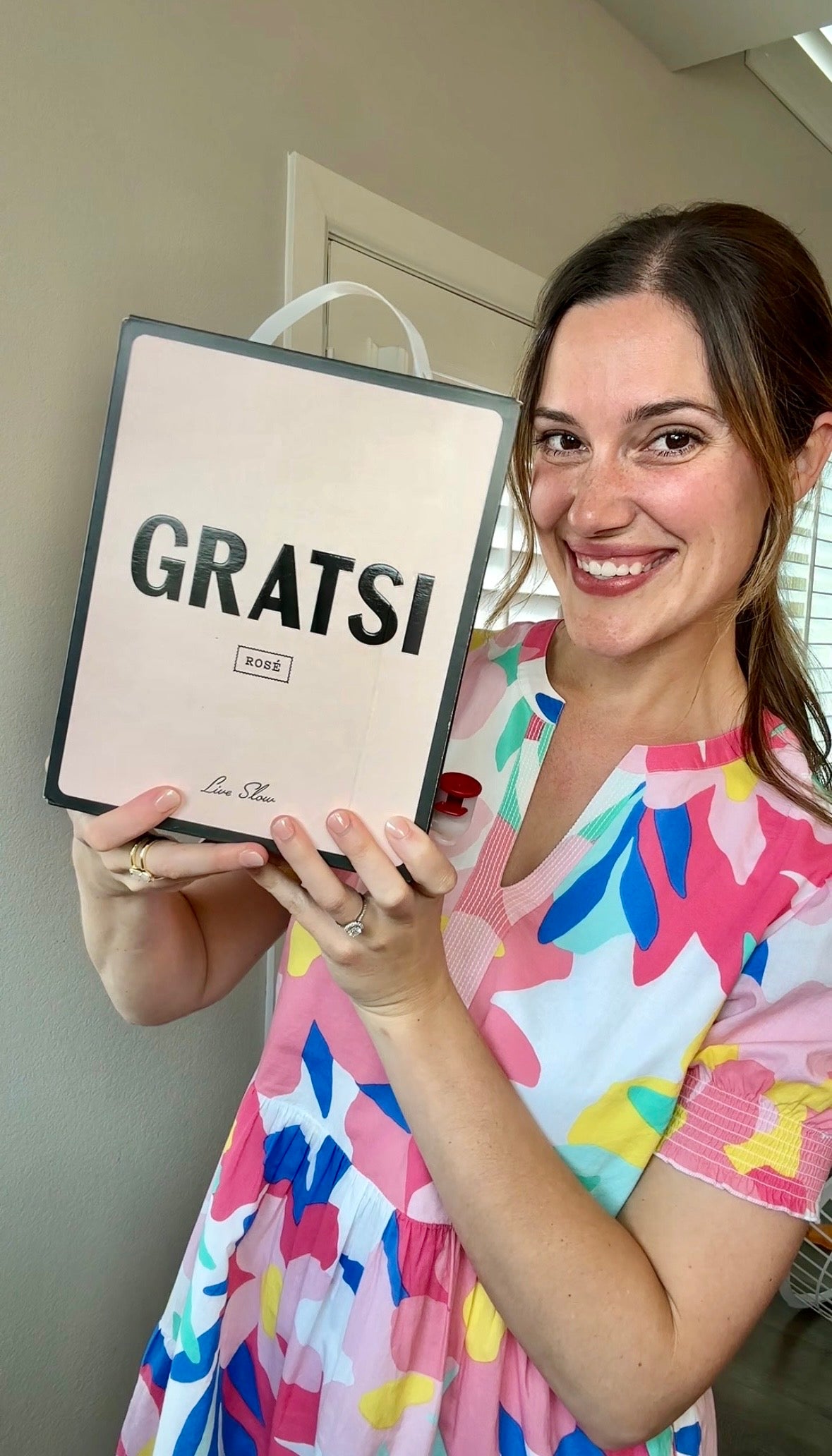 A smiling woman in a colorful floral dress holds up a box labeled GRATSI ROSÉ with both hands. She stands indoors near a window with blinds, and the box has a light pink background.