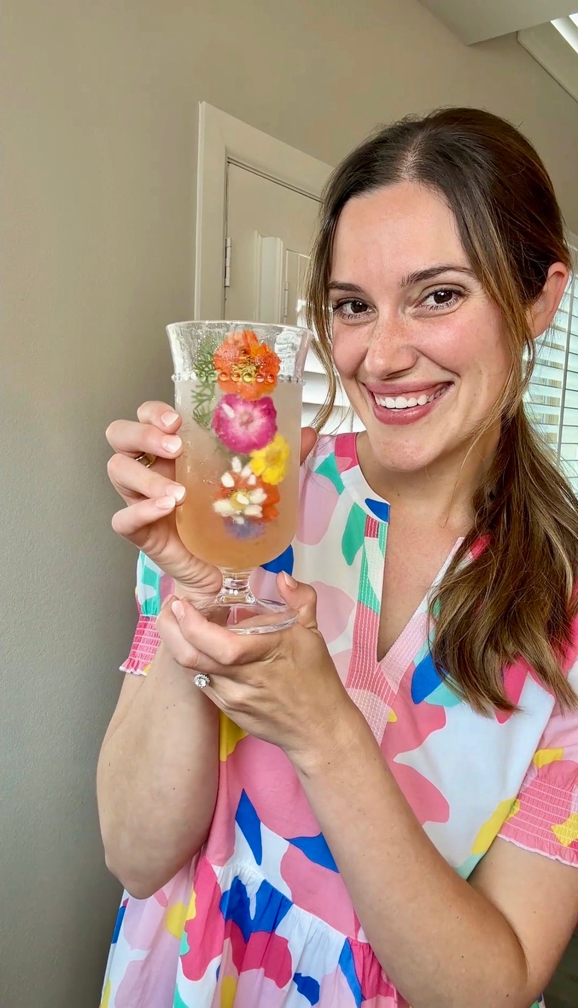 A smiling woman in a colorful dress holds up a clear glass of pink drink filled with ice cubes containing colorful edible flowers and herbs, standing indoors near a window.