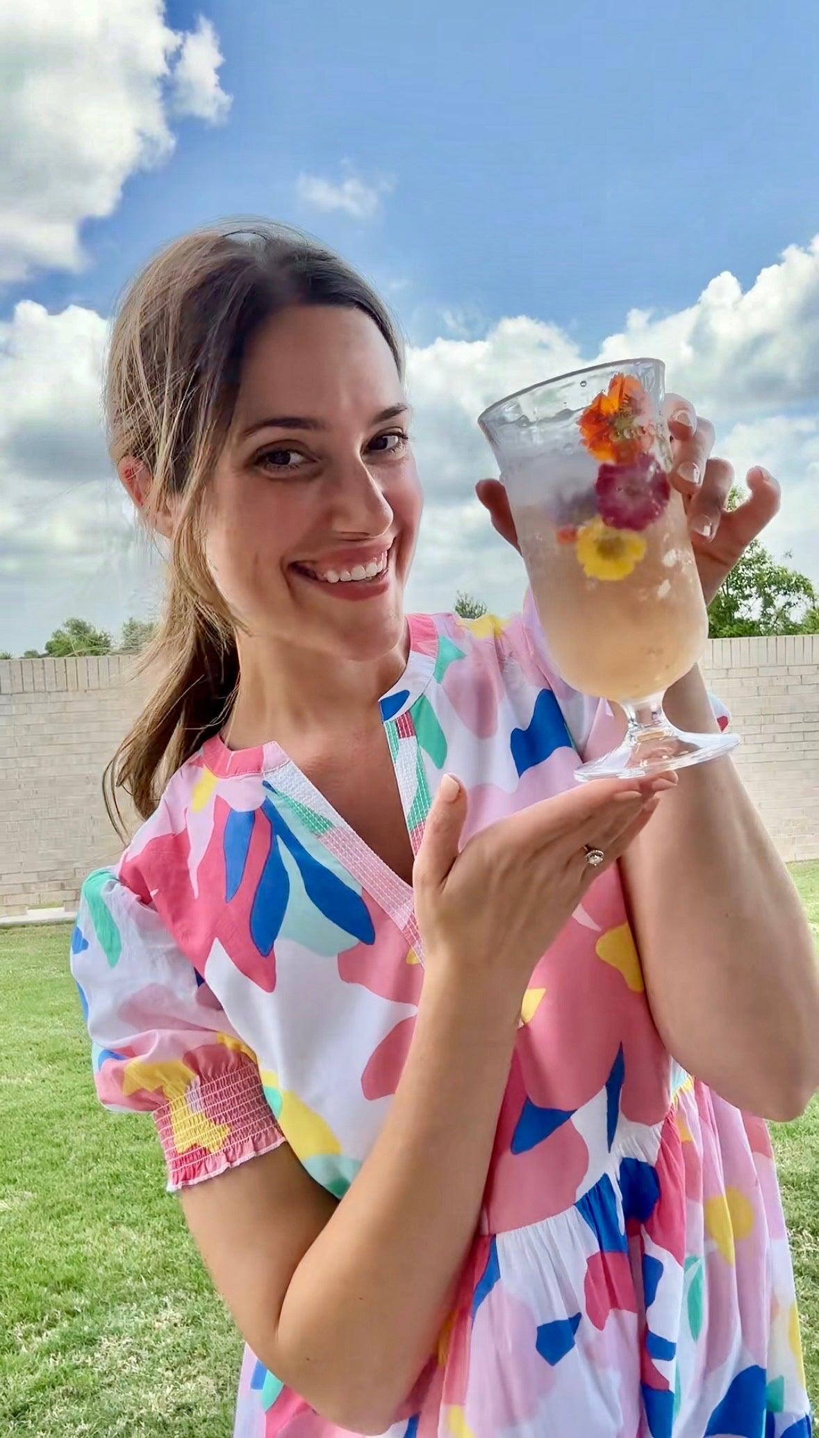 A woman wearing a colorful floral dress smiles and holds up a glass of iced beverage with floating edible flowers, standing outside on a grassy lawn with a brick wall and blue sky in the background.