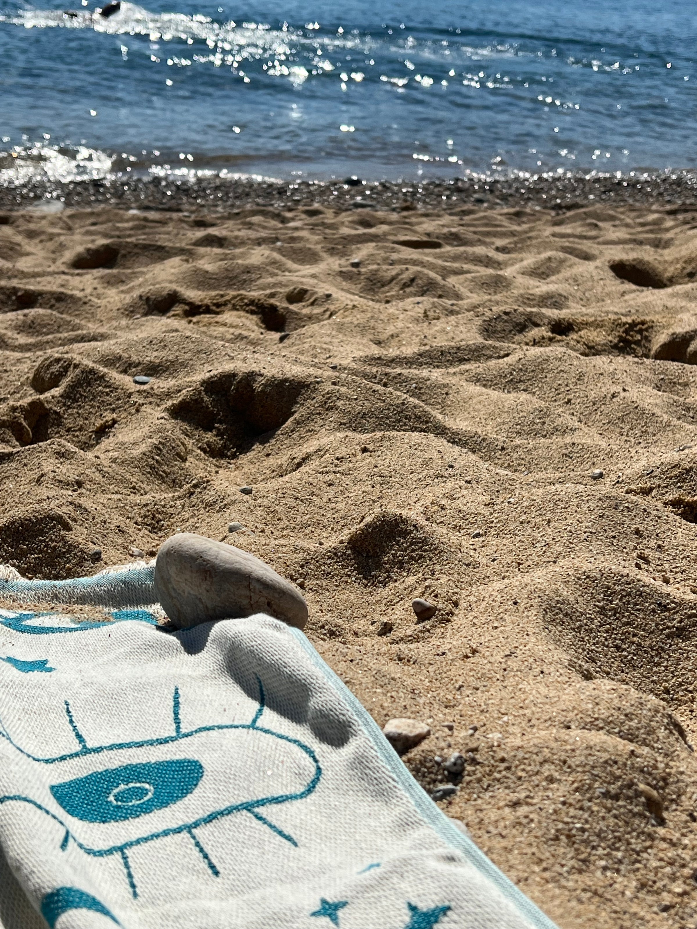 A beach scene with golden sand, a white and blue towel partially visible in the foreground, a small smooth stone on the towel, and waves gently lapping the shore in the background.