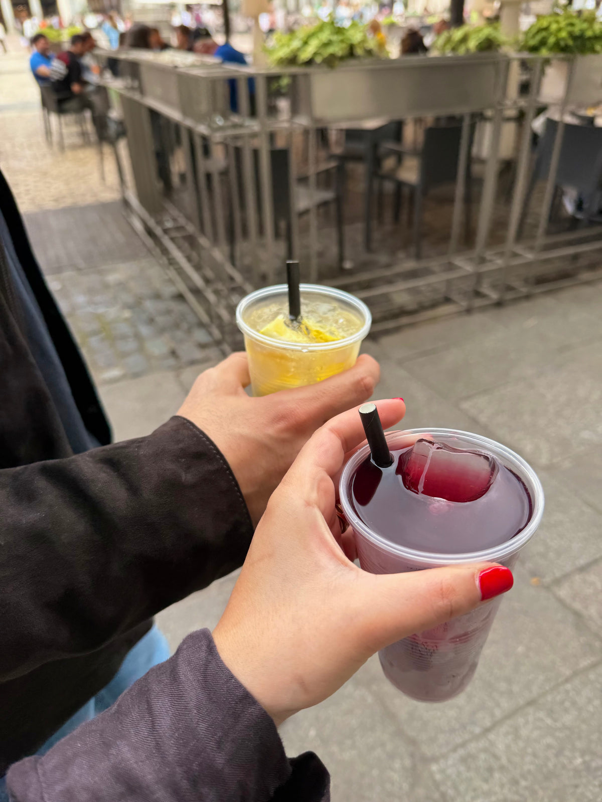 Two people holding plastic cups with drinks—one yellow, one purple—standing outdoors by metal railings. One hand has red nail polish; people and tables are visible in the blurred background.