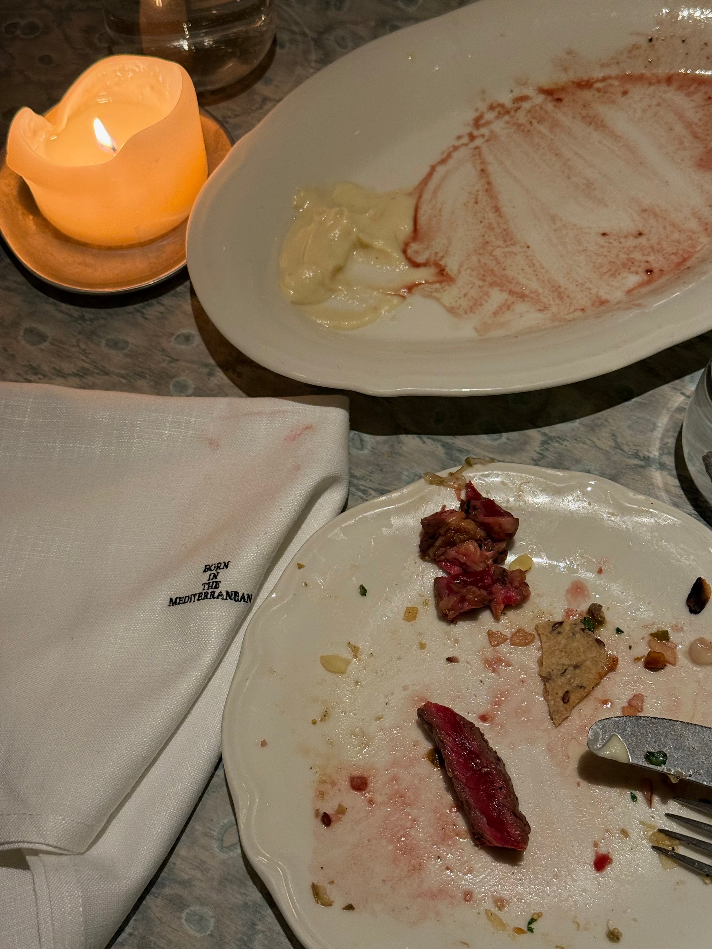 A nearly empty dinner plate with a few pieces of food, a used fork, a napkin with New York MEDITERRANEAN embroidered, and a melted candle on a table with dim lighting.