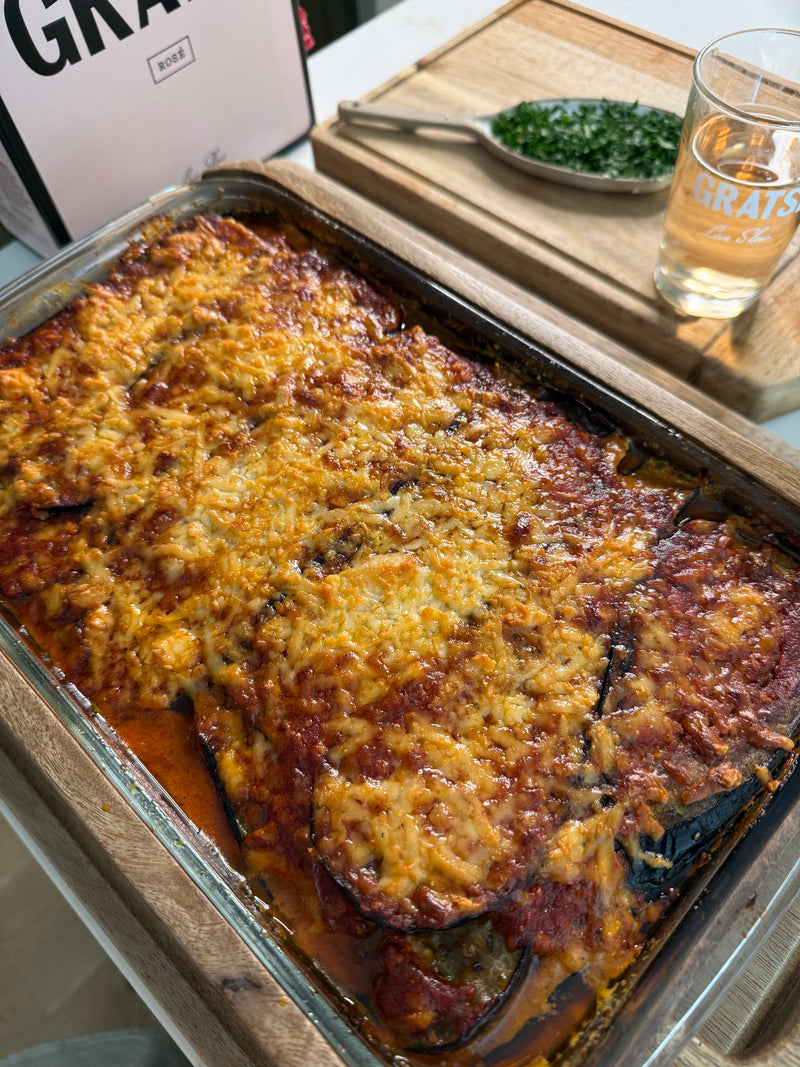 A glass baking dish filled with baked eggplant parmesan topped with melted cheese and tomato sauce, sitting on a wooden tray. A drink and a small dish of chopped herbs are in the background.