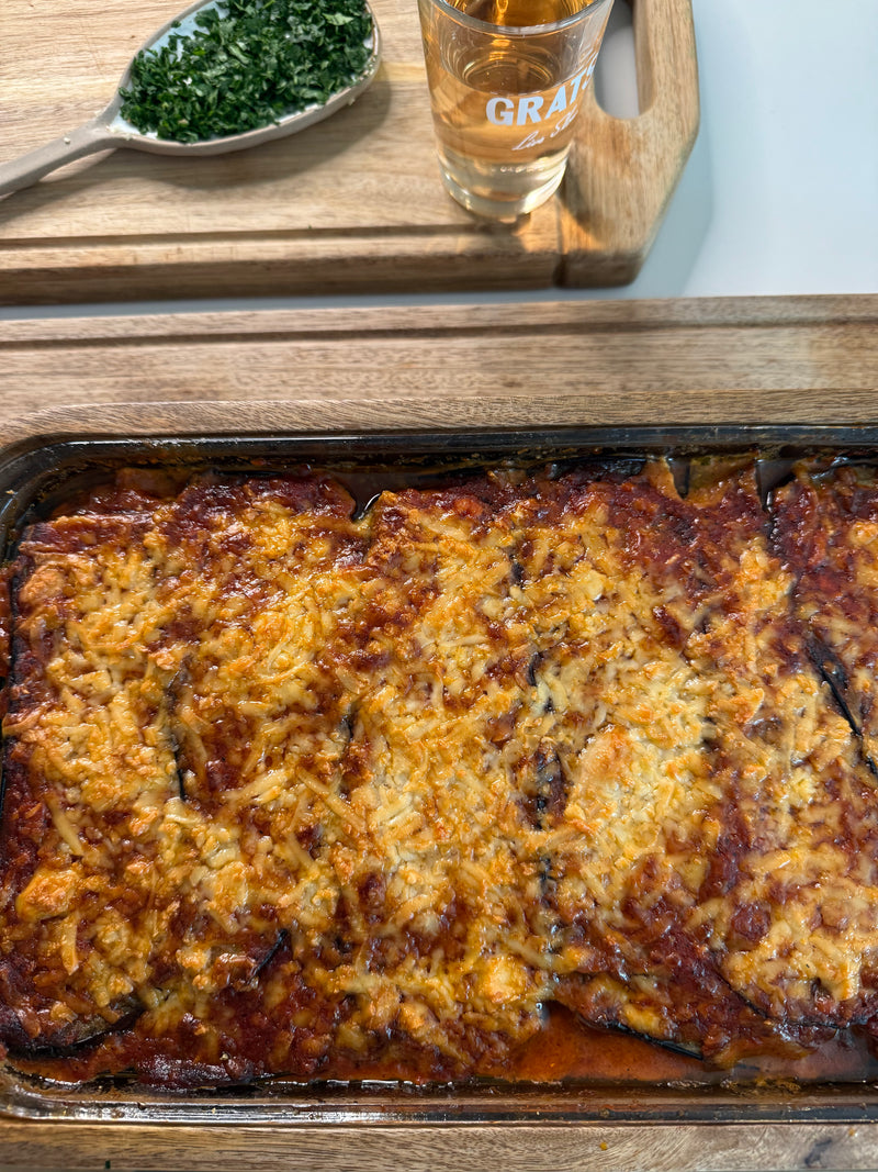 A freshly baked casserole with a golden, cheesy top sits on a wooden surface. Nearby, a glass of a light beverage and a spoonful of chopped herbs rest on a wooden cutting board.