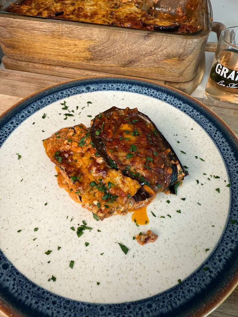 A serving of moussaka topped with herbs on a white plate with a blue rim. In the background, a glass baking dish with more moussaka and a drink in a glass are visible.