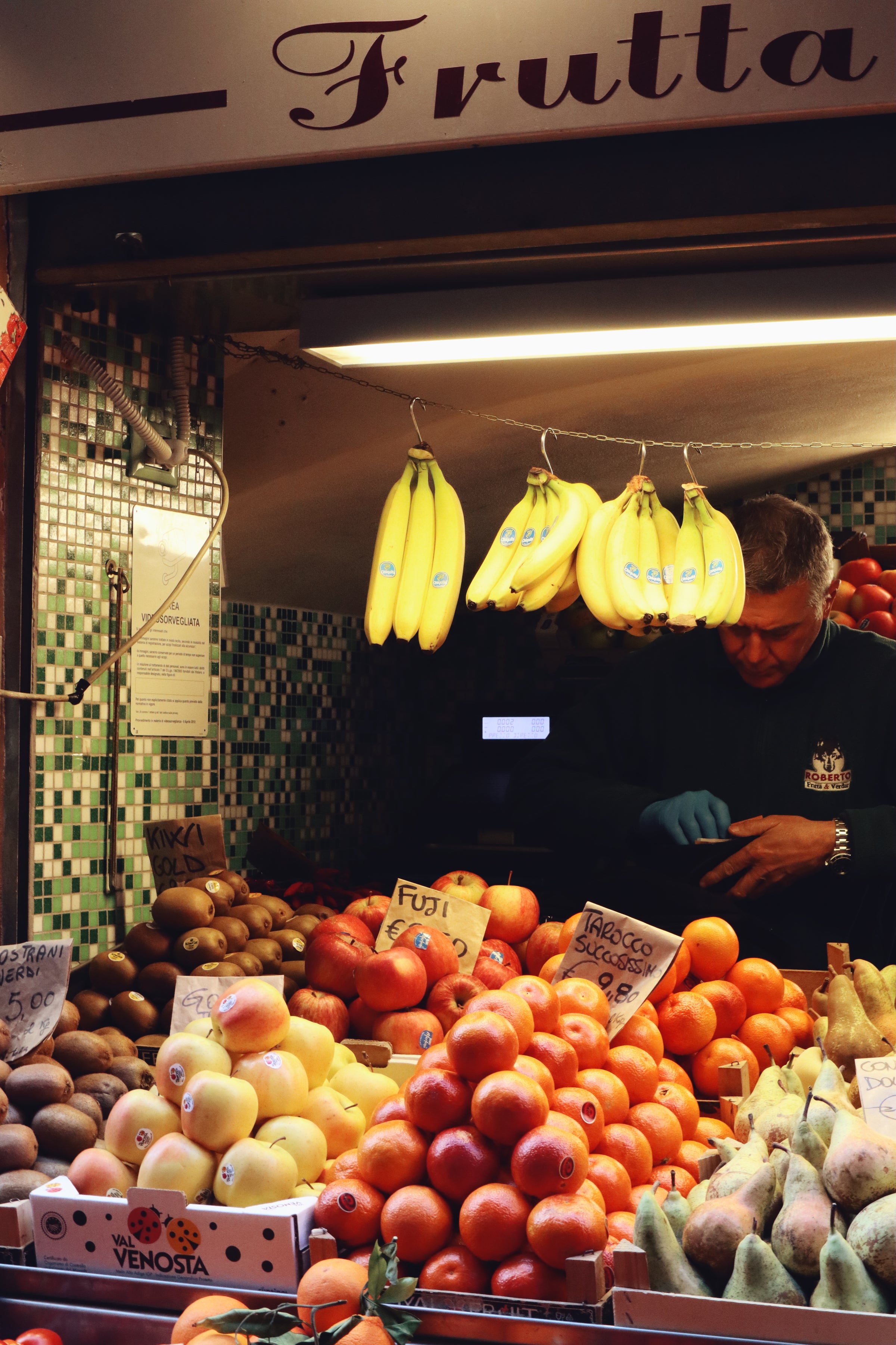 A man stands behind a colorful fruit stall with bananas, apples, pears, oranges, and kiwis displayed. Handwritten signs label each type of fruit. The stall has a tiled wall and a Frutta sign above.