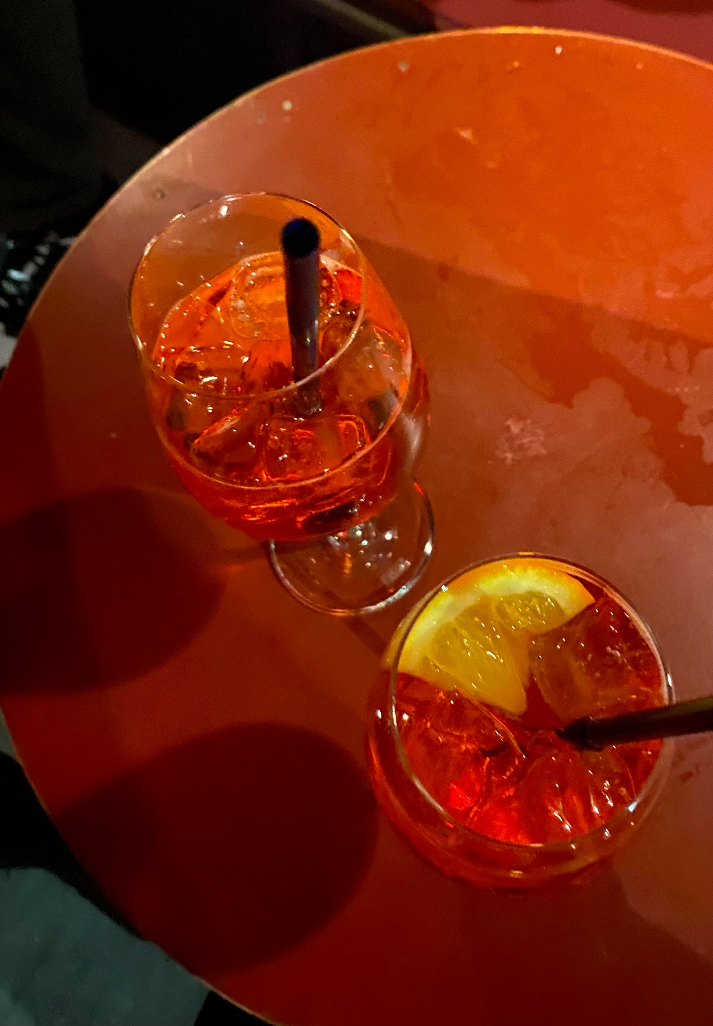Two glasses with orange-colored drinks and ice sit on a round, red table. One glass has a slice of orange and a straw; the other has a straw and no fruit garnish.