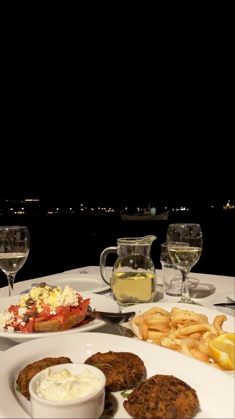 A nighttime outdoor dining scene with two glasses of white wine, a jug of water, falafel, fries, bread with tomatoes and feta, and city lights visible in the distance.