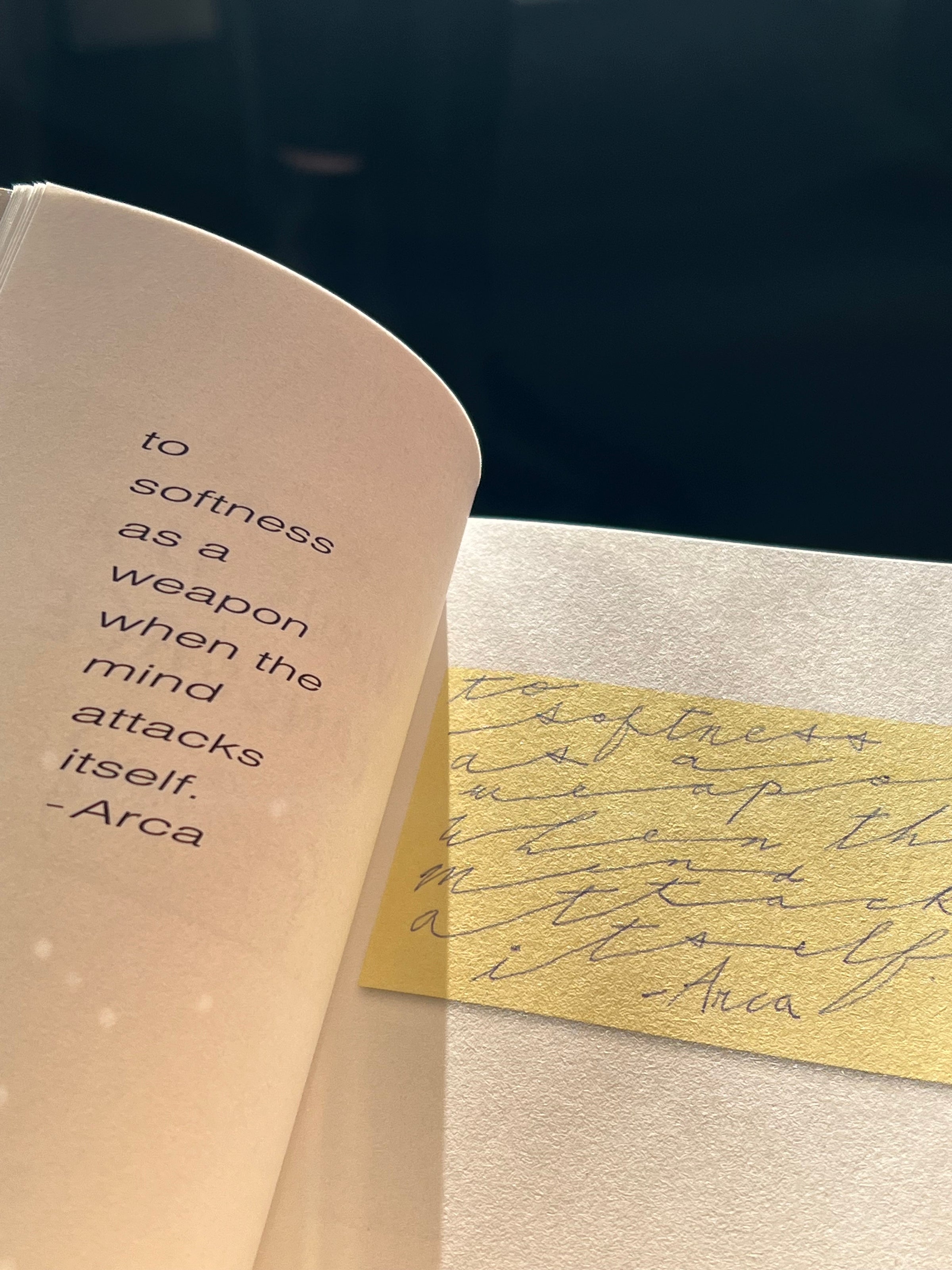 A book open to a page with the text to softness as a weapon when the mind attacks itself. - Arca alongside a yellow handwritten note, lit by sunlight.