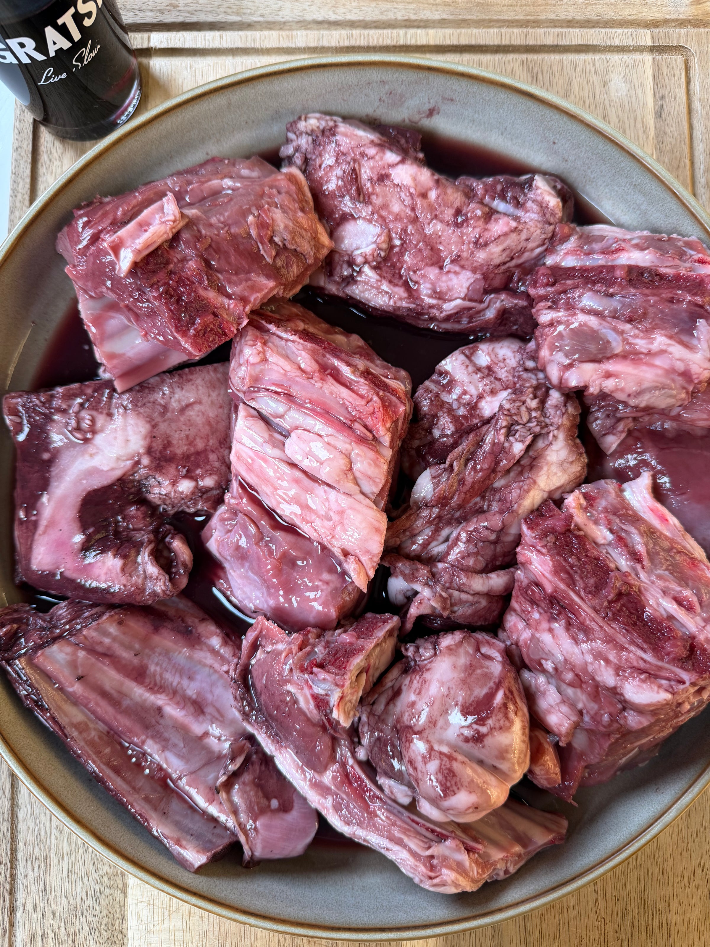 A bowl filled with several raw beef short ribs soaking in a red marinade or liquid, placed on a wooden surface. A can of beer is partially visible in the top left corner.