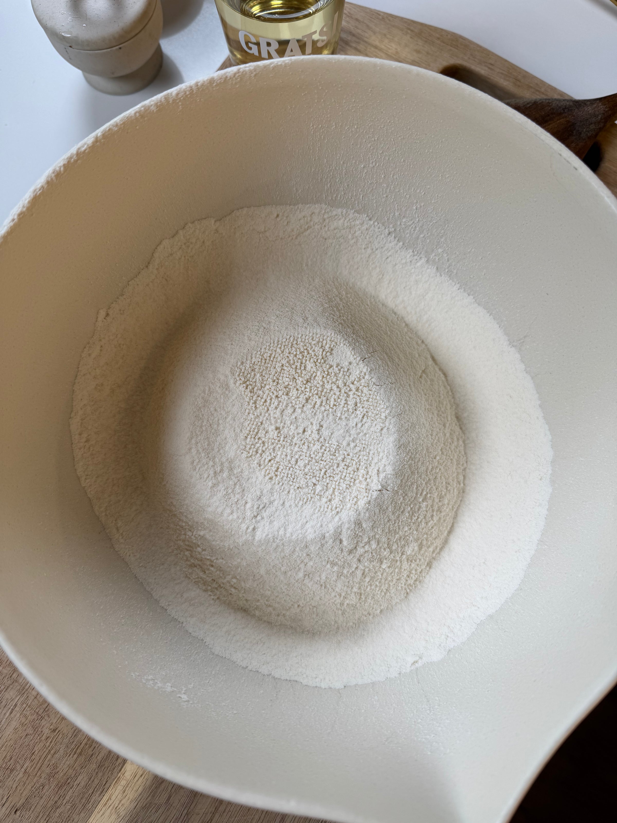A white mixing bowl filled with flour and active dry yeast, with a wooden spoon and a can of grated cheese visible in the background on a wooden surface.
