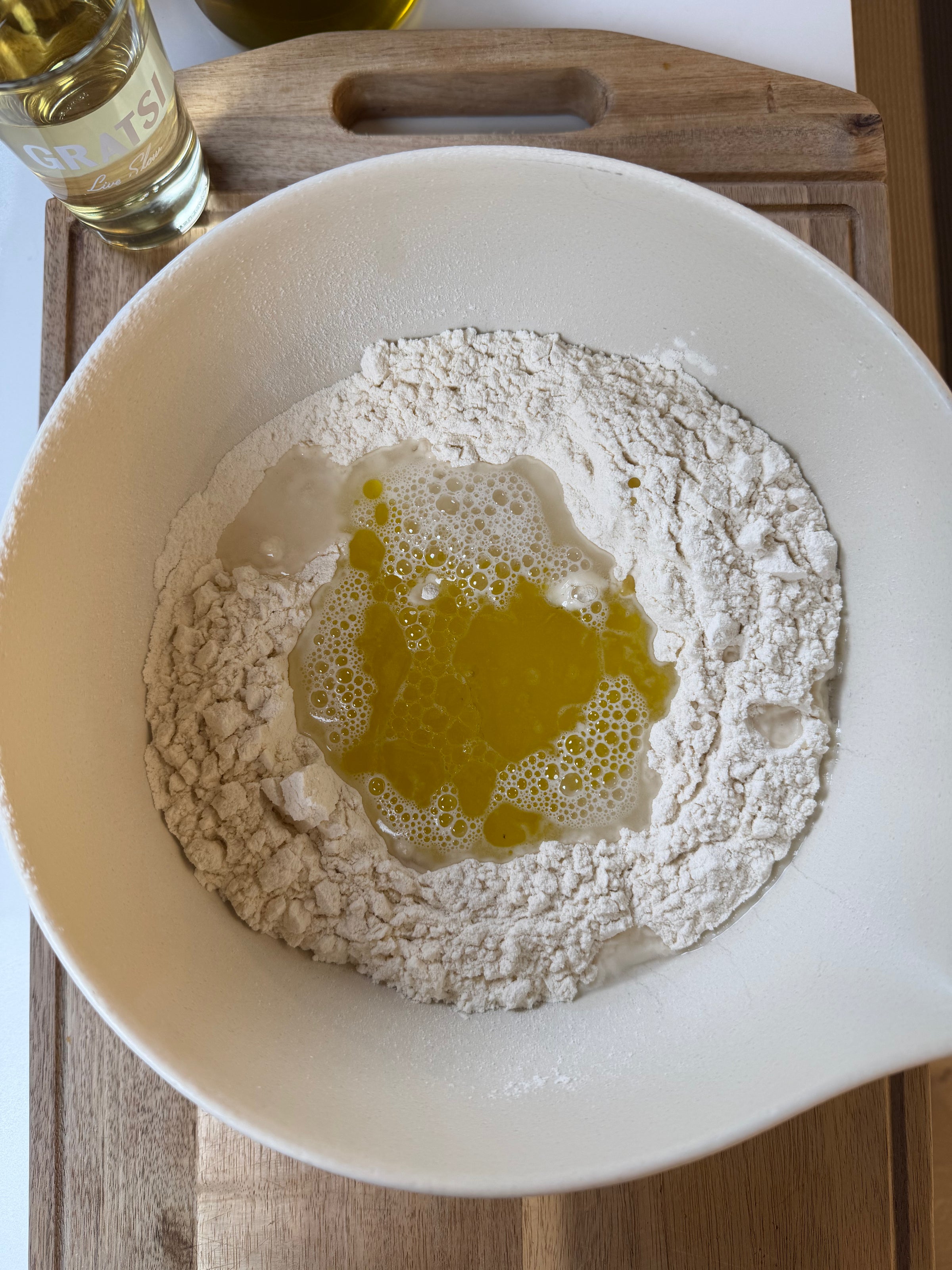 A white bowl with flour, water, and olive oil sits on a wooden cutting board. A bottle of olive oil and part of a glass are visible beside the bowl.