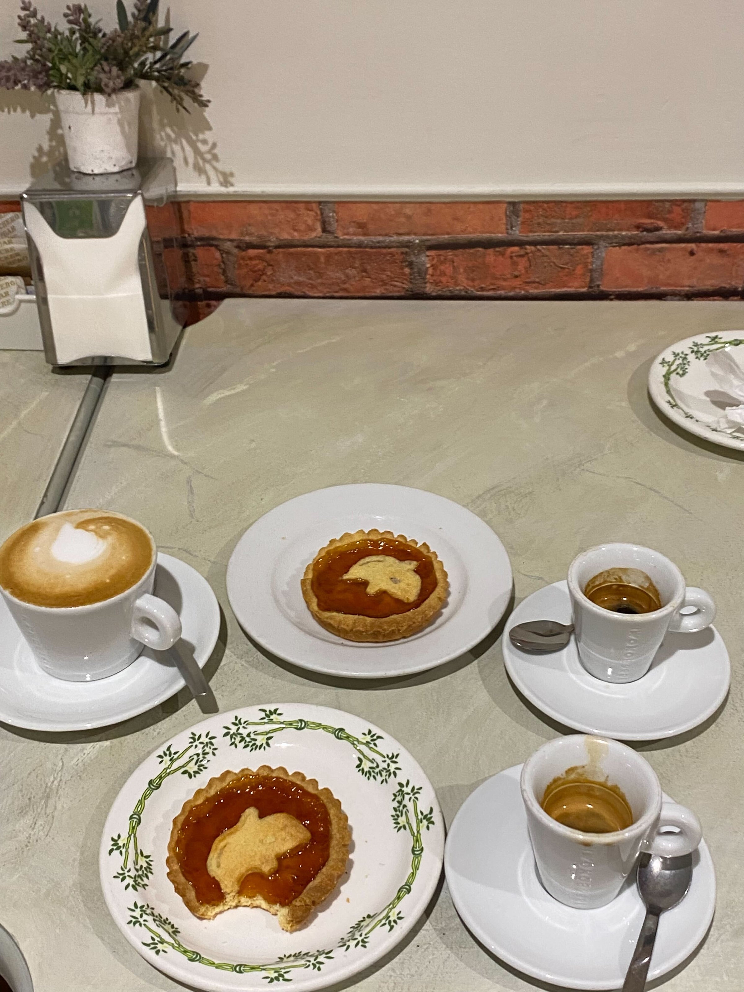 Three cups of coffee, two espressos and a cappuccino, are on a marble table with two jam tarts on plates. One tart has a bite taken out. A plant and a napkin holder sit in the background near a brick wall.