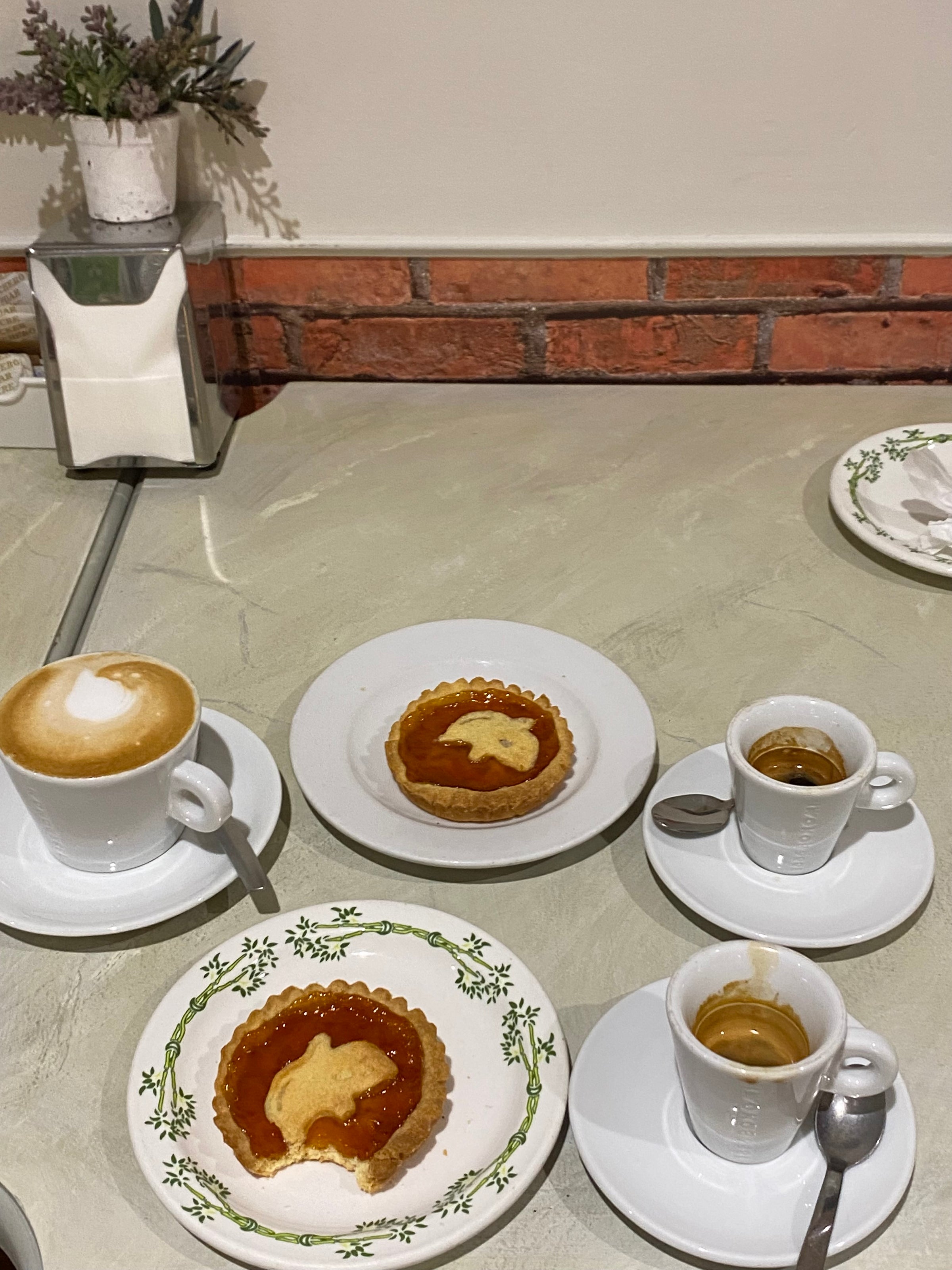 Three small white cups of coffee sit beside two jam tarts on plates, one with a bite taken out, on a table with a brick wall and a small plant in the background.