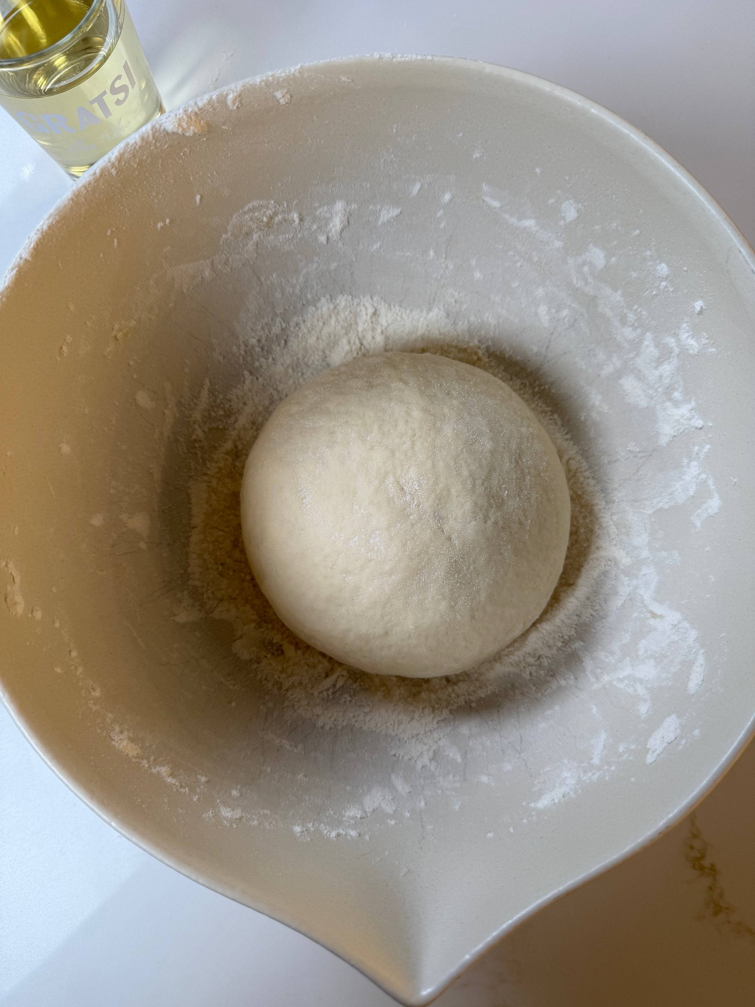 A round ball of dough sits in a large, white mixing bowl dusted with flour. A small glass of yellow liquid, likely oil, is partly visible in the top left corner.