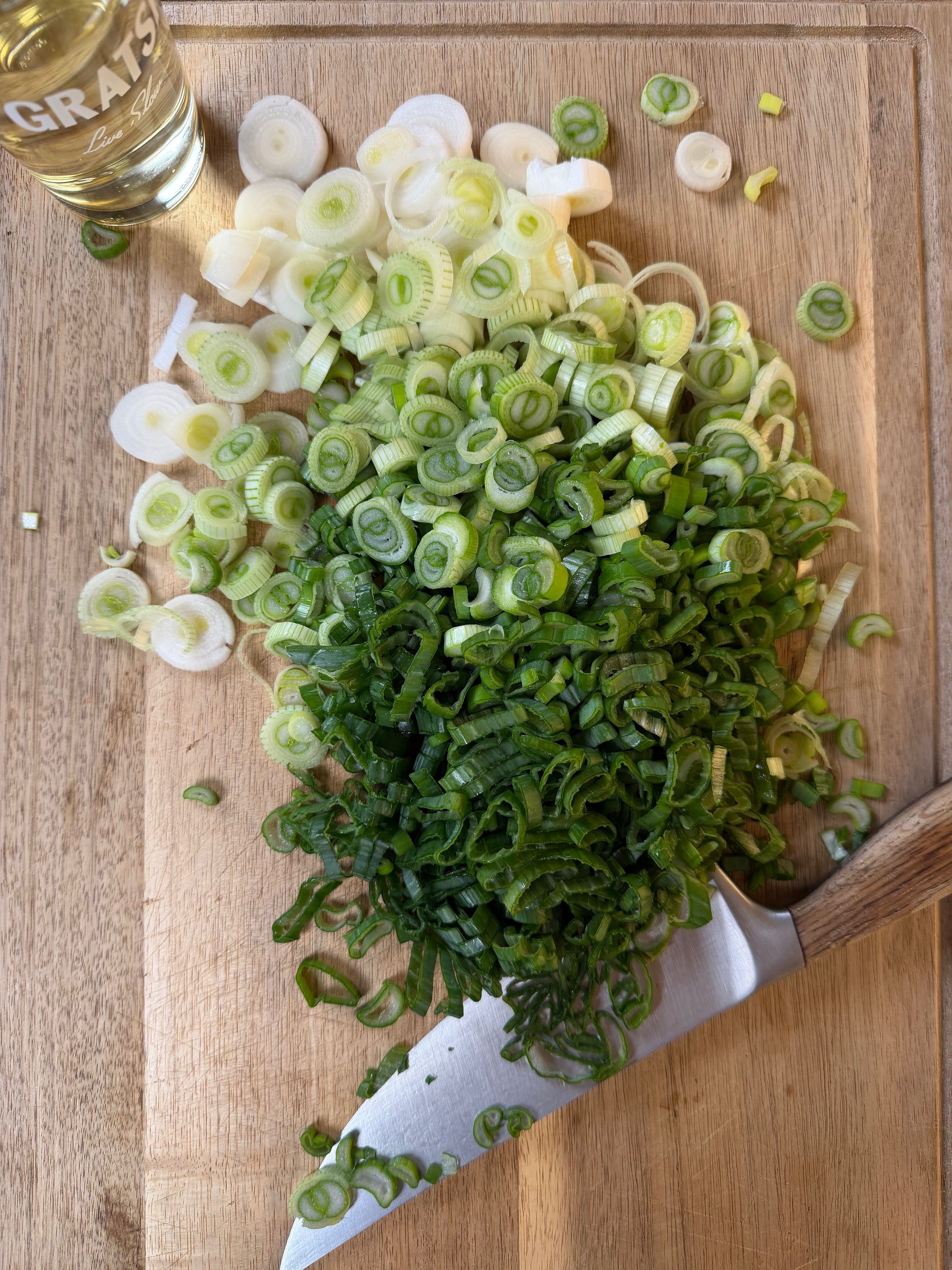 A pile of chopped green onions, both white and green parts, sits on a wooden cutting board next to a knife. A bottle labeled GRATS is partially visible in the top left corner.