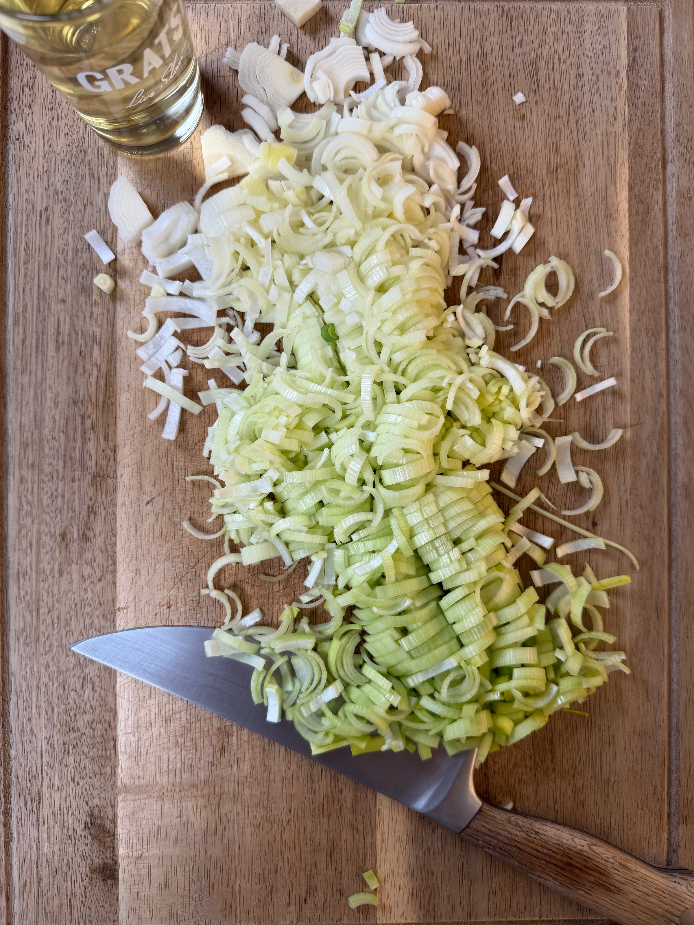 A pile of sliced leeks on a wooden cutting board, next to a chefs knife. A glass of white wine sits in the upper left corner of the board.