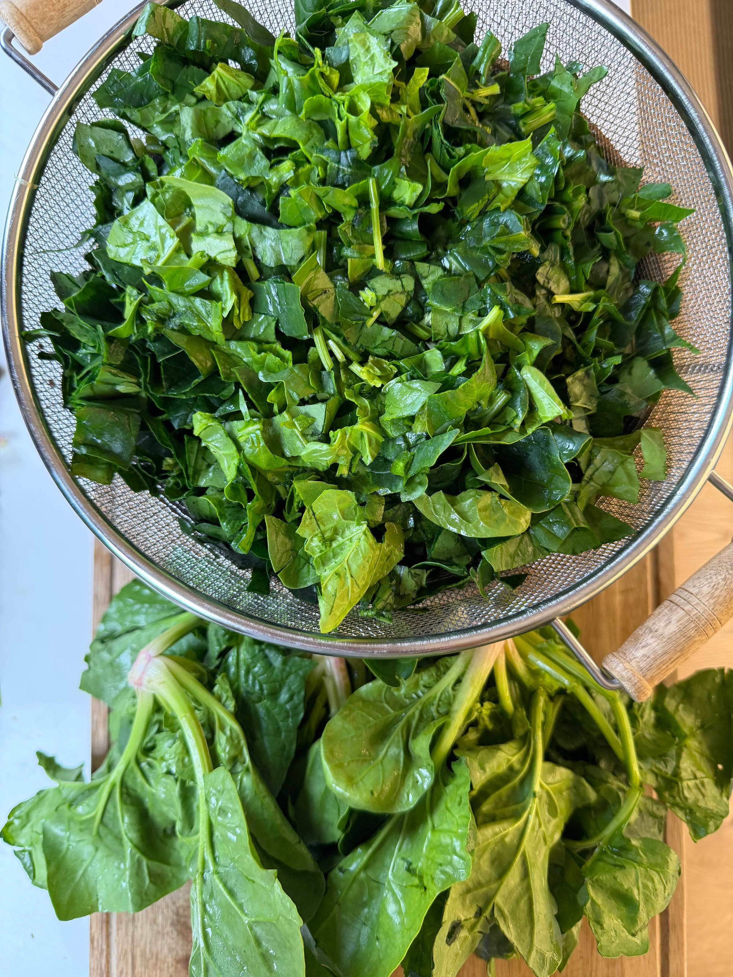 A metal strainer filled with chopped spinach sits on a wooden surface, with fresh, unchopped spinach leaves lying next to it.