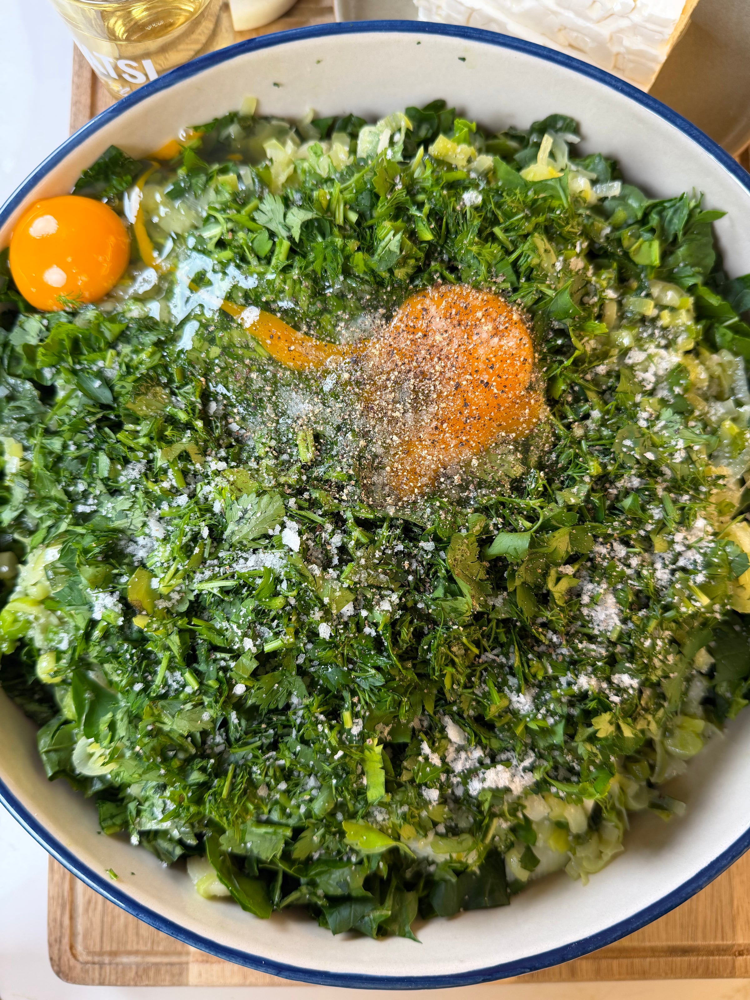 A bowl filled with chopped greens, fresh herbs, an egg yolk, spices, and salt, ready to be mixed together, sits on a wooden surface.