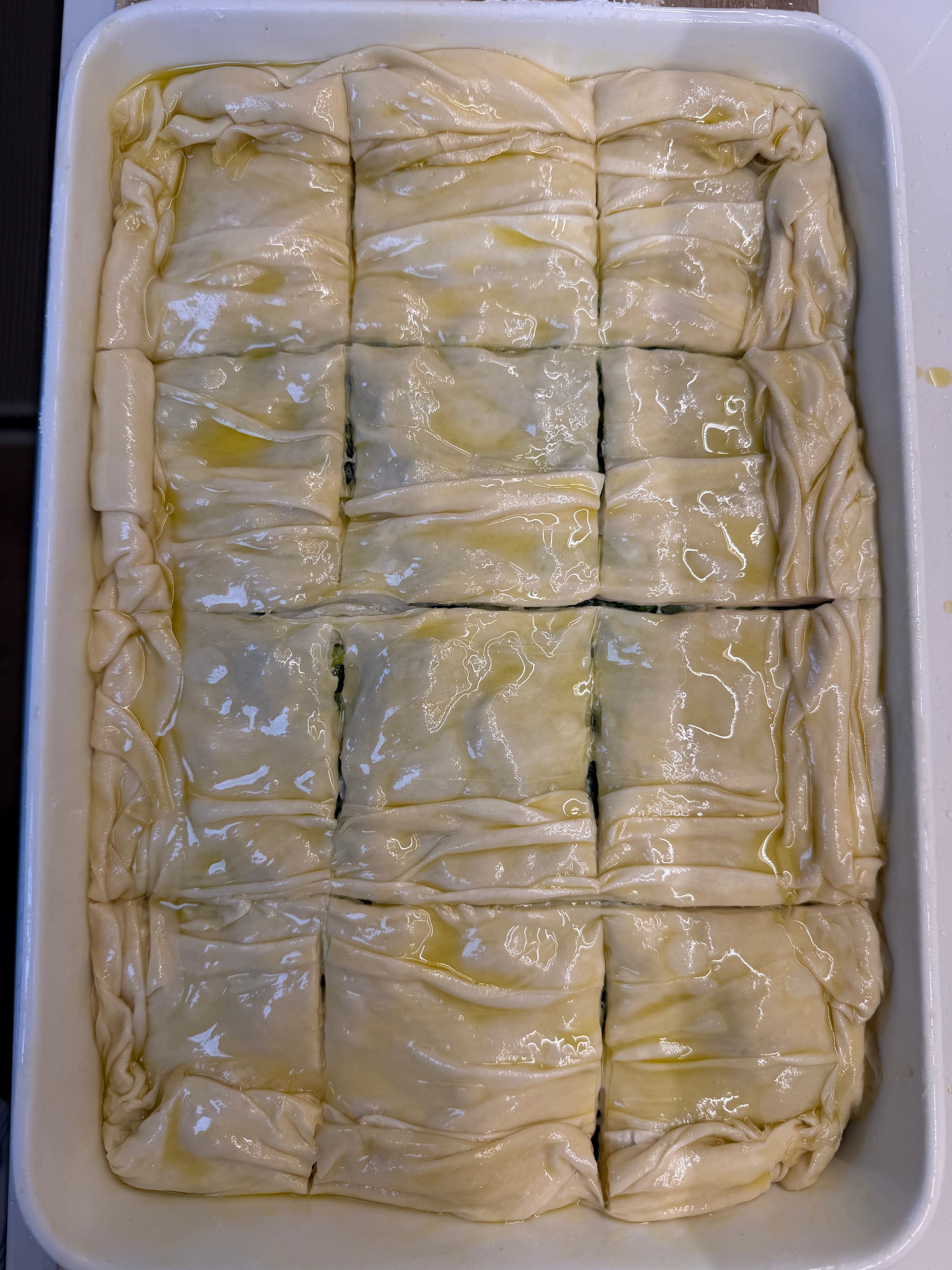 A rectangular baking dish filled with unbaked, golden oiled phyllo dough, scored into twelve portions, ready for baking. The surface is shiny and slightly wrinkled.