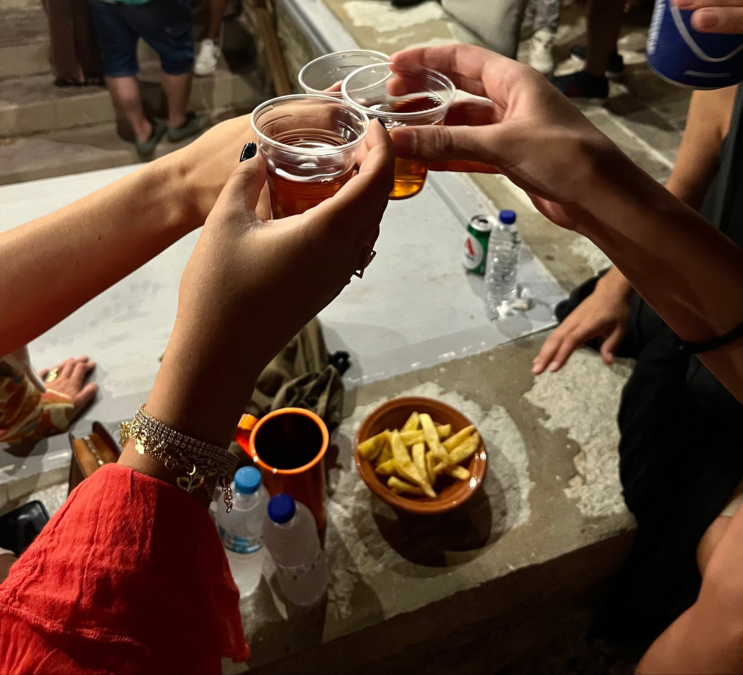 Three people clink plastic cups with drinks, seen from above. A bowl of fries, water bottles, and personal items are on a stone surface nearby. The scene appears to be at an outdoor gathering at night.