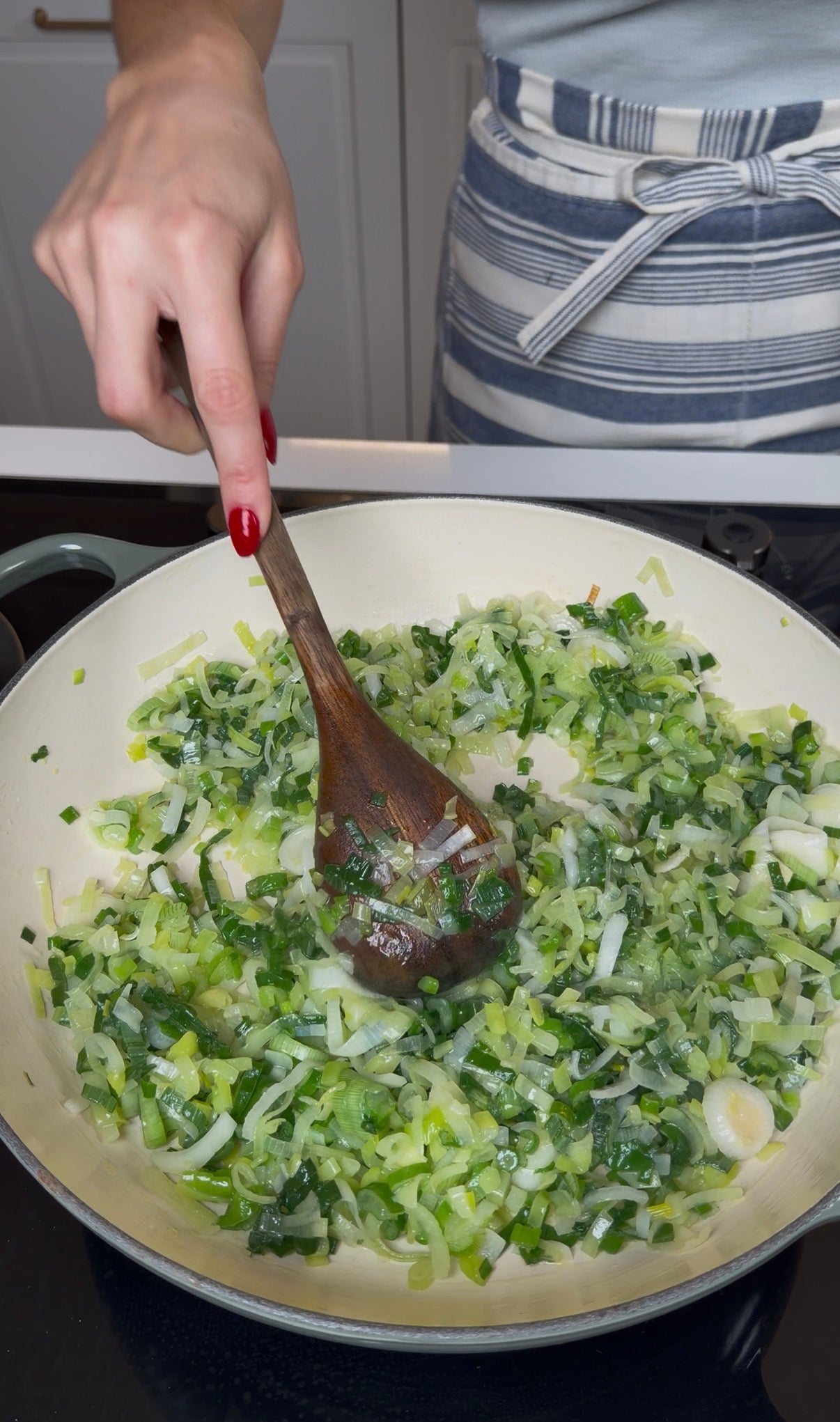A person in a striped apron stirs chopped leeks and onions in a white pan with a wooden spoon, cooking the vegetables on a stovetop.