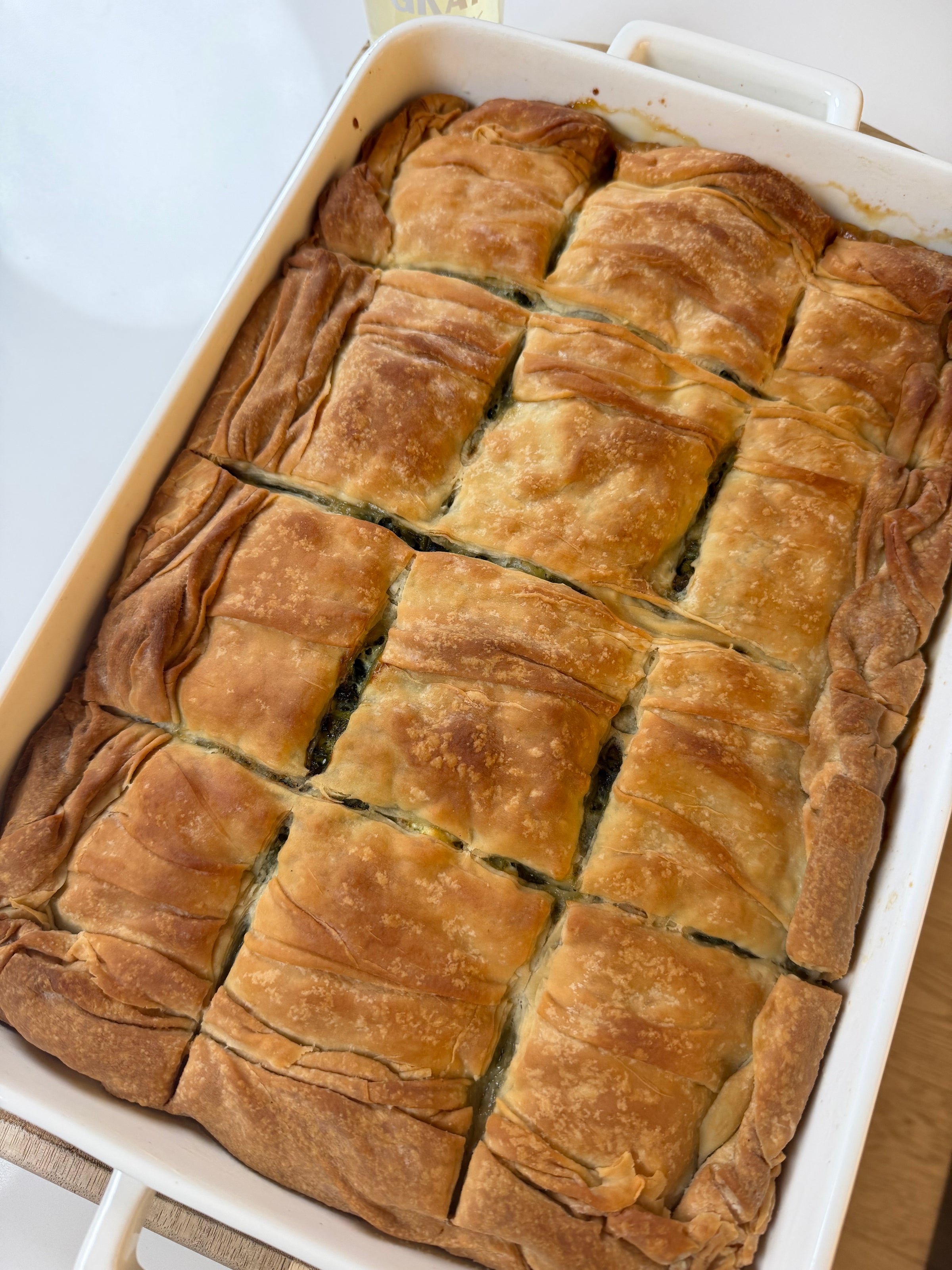 A golden-brown baked pastry, cut into twelve rectangular pieces, sits in a white rectangular baking dish. The pastry appears flaky and has visible layers.