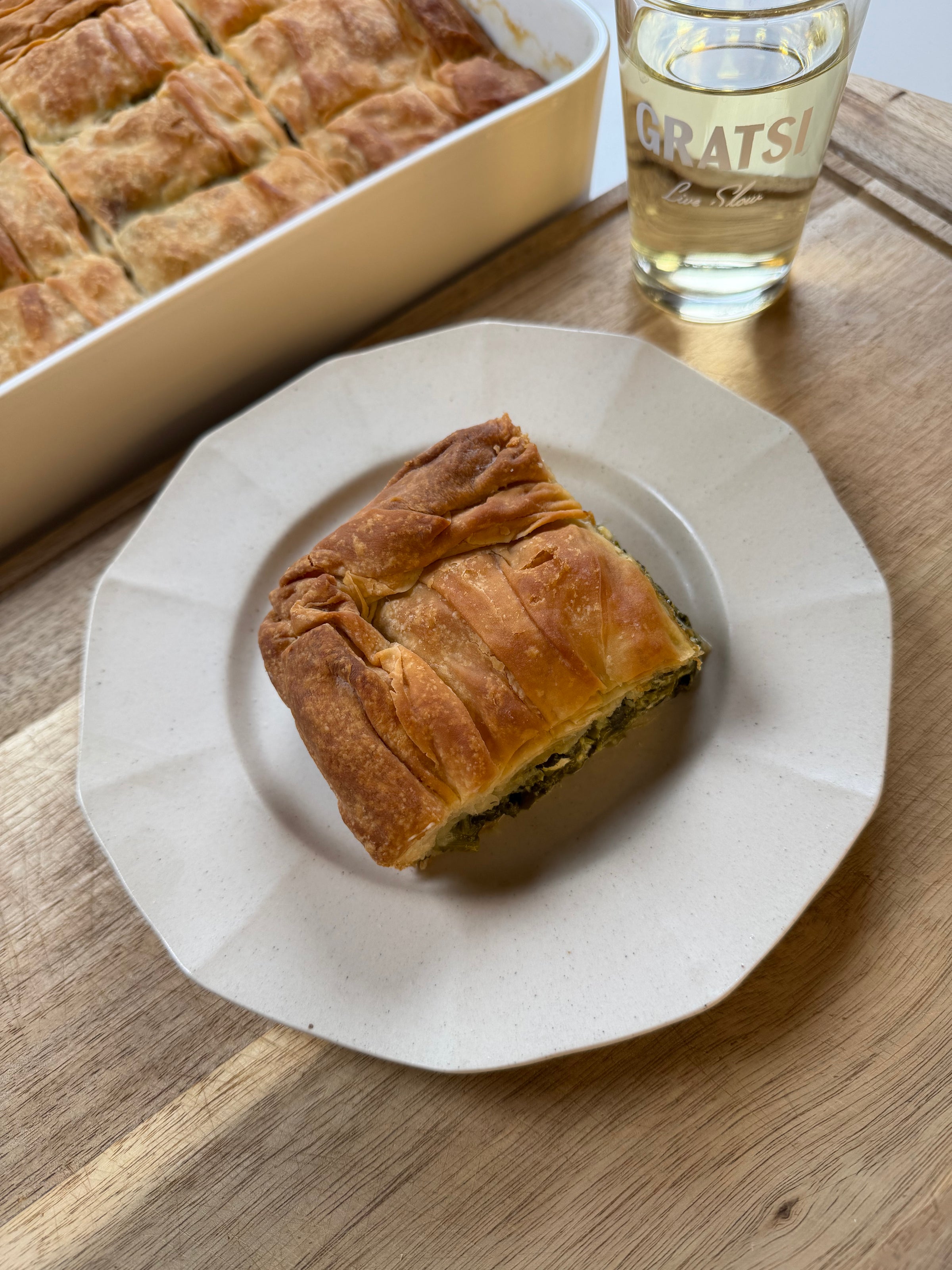 A slice of baked savory pie, likely spinach and cheese, sits on a white plate next to a glass of white wine and a baking dish with more pie, all on a wooden table.