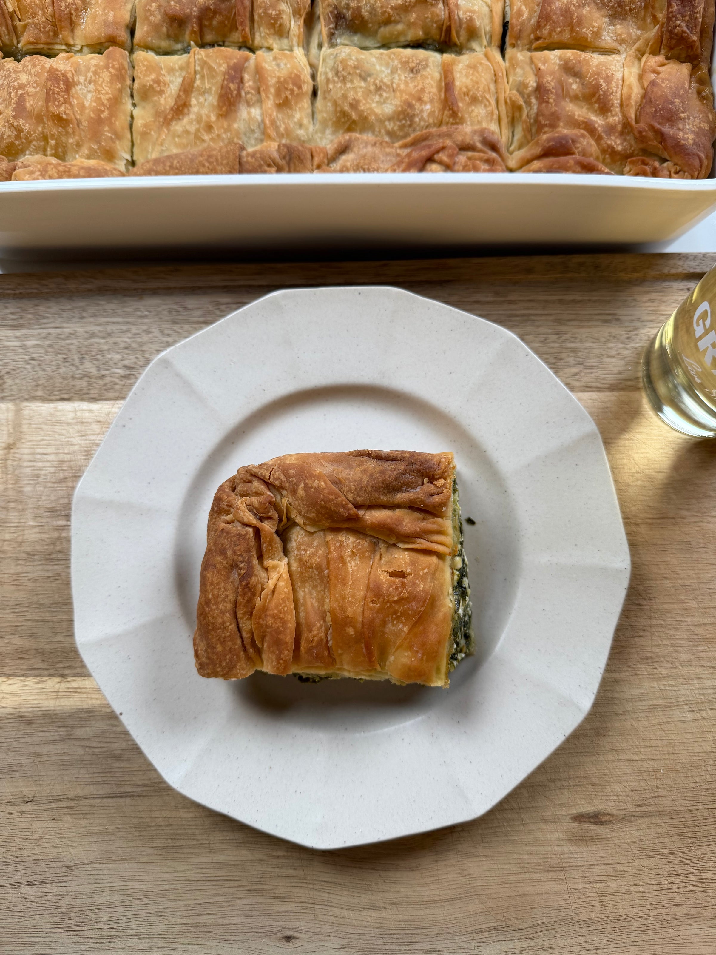 A square piece of golden, flaky spinach pie is served on a white plate. In the background, a larger dish of the same pie is visible, along with a glass of clear liquid on a wooden surface.