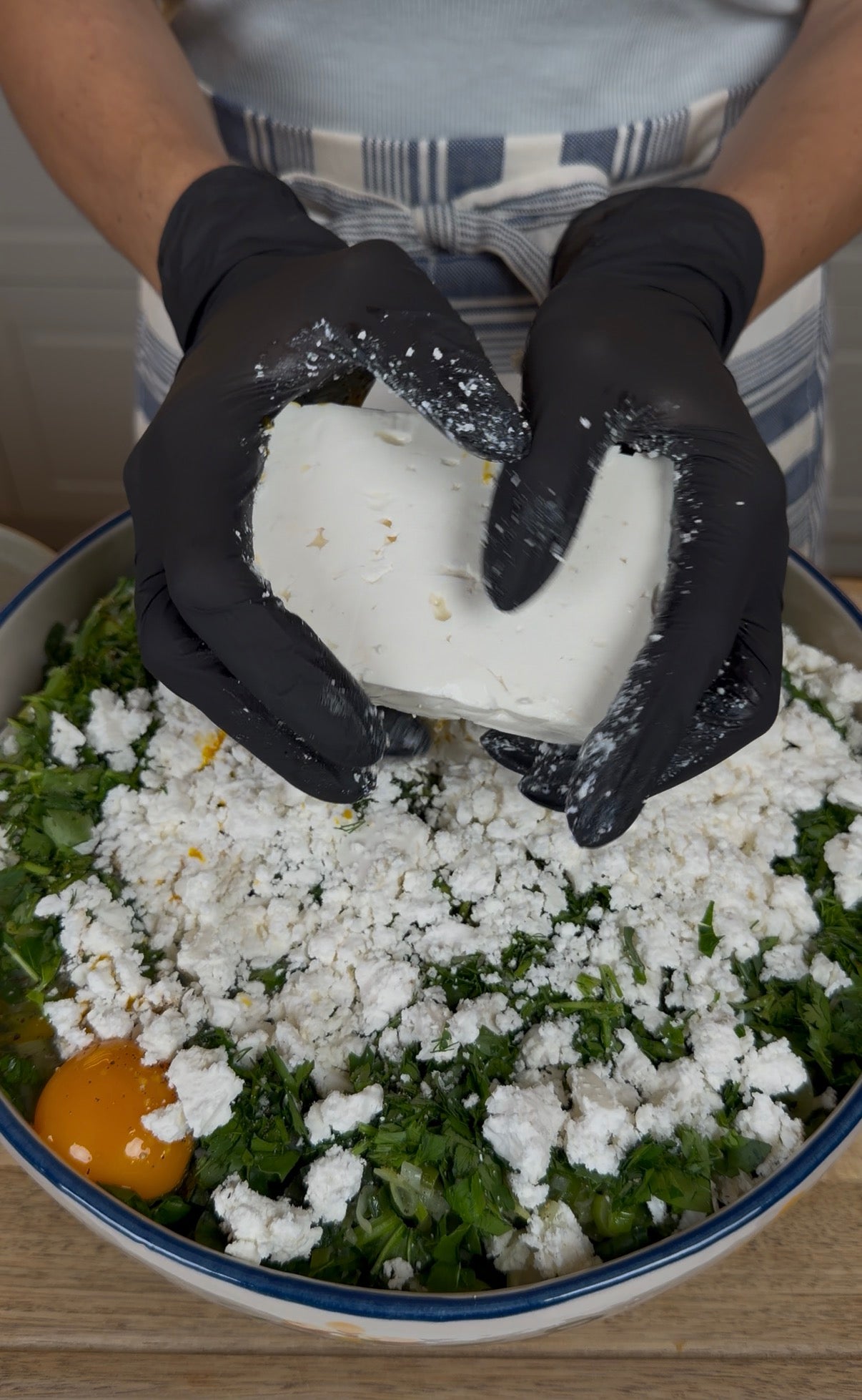 A person wearing black gloves crumbles a block of white cheese over a bowl filled with chopped greens and an egg.
