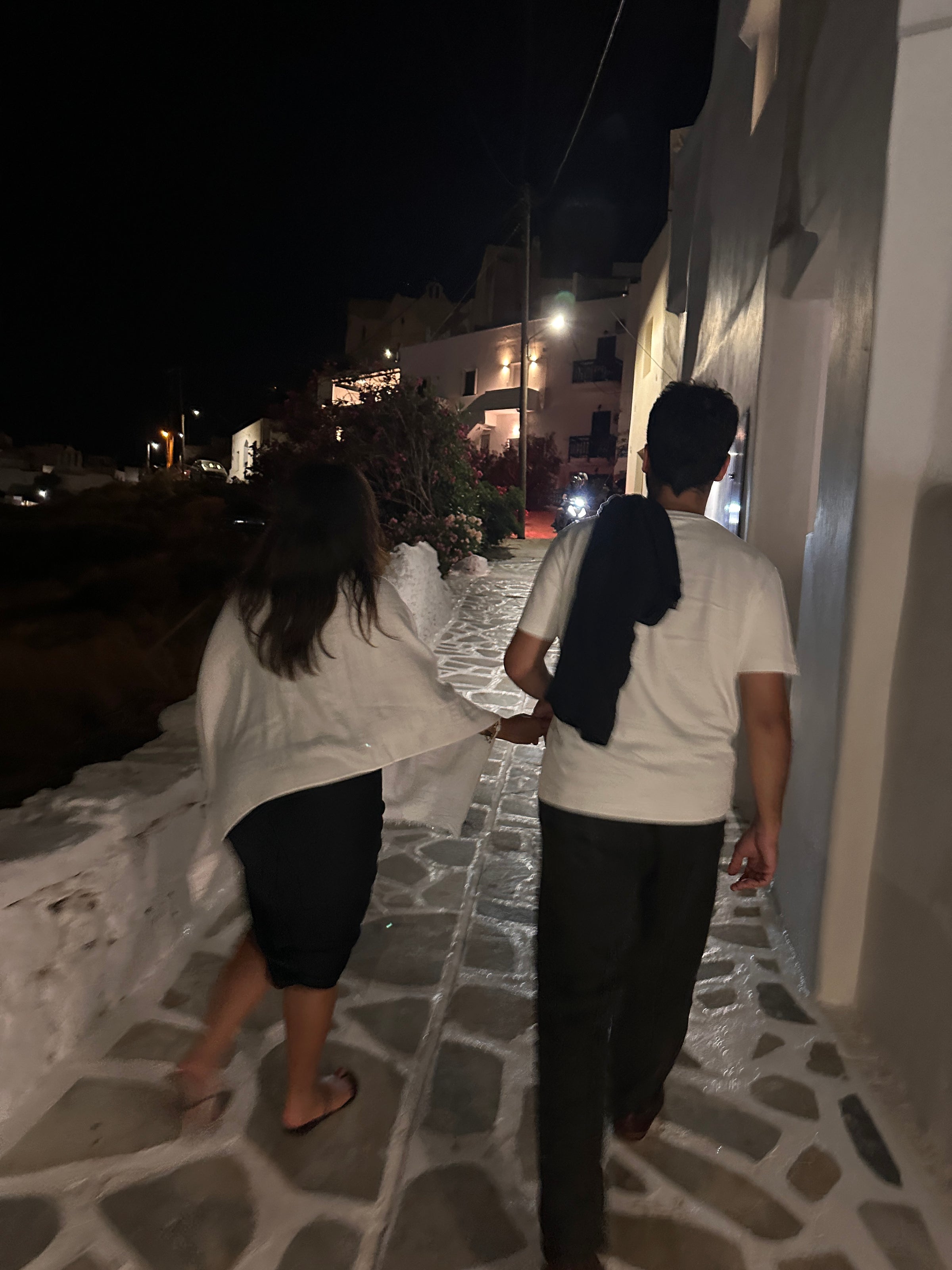 Two people walk side by side on a stone path at night, near white buildings. One drapes a sweater over their shoulders, and the path is illuminated by lights along the street.