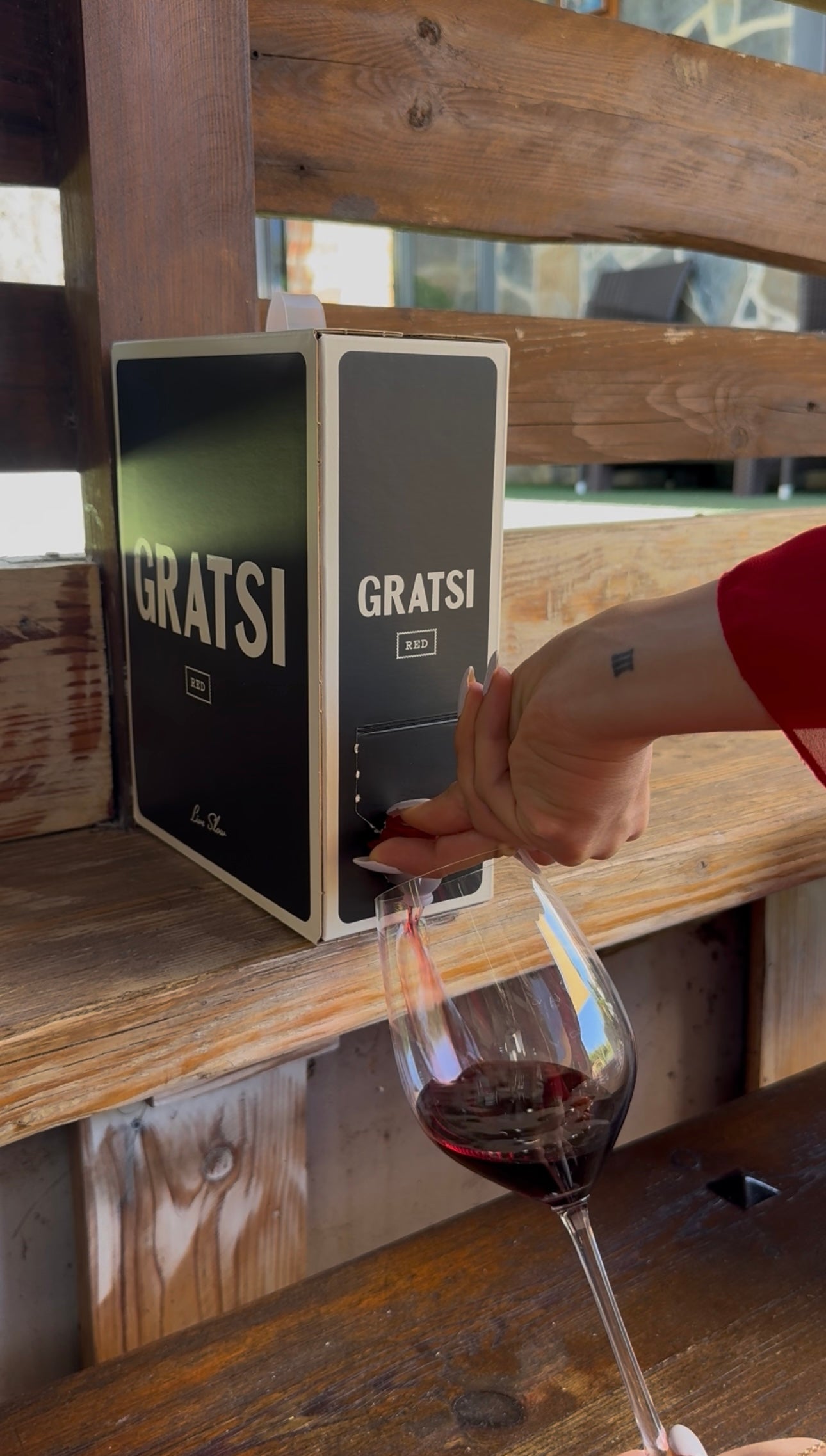 A person in a red sleeve fills a wine glass with red wine from a black and white GRATS! boxed wine dispenser on a wooden shelf.