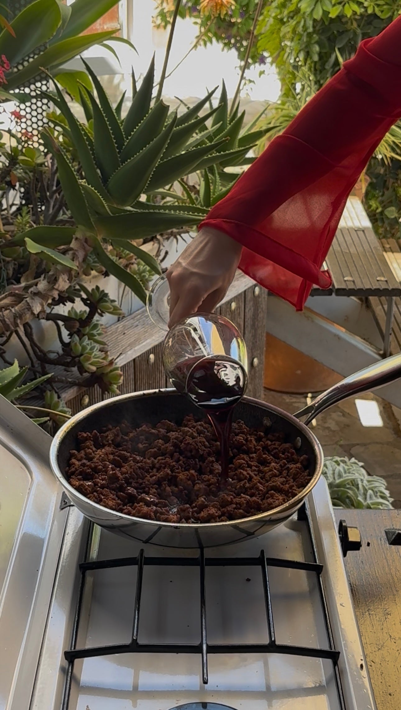 A person in a red sleeve pours red wine from a glass into a frying pan filled with cooked ground meat on a stove, with green plants visible in the background.