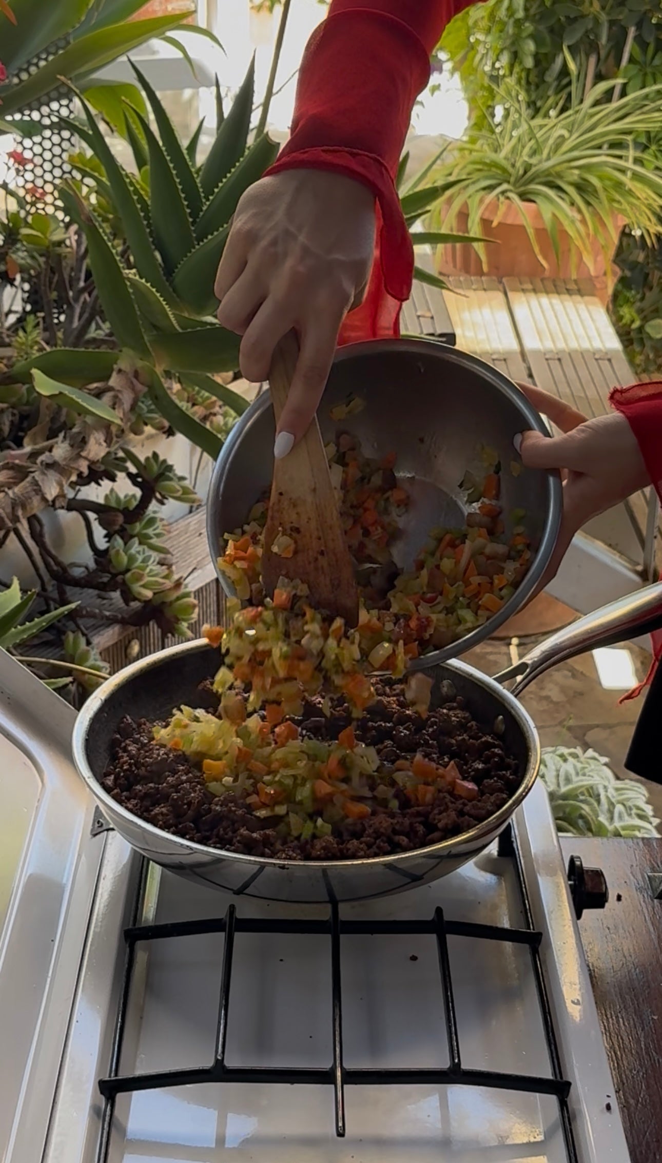 A person pours chopped vegetables from a bowl into a pan with cooked food on a stove, surrounded by lush green plants in an outdoor setting.
