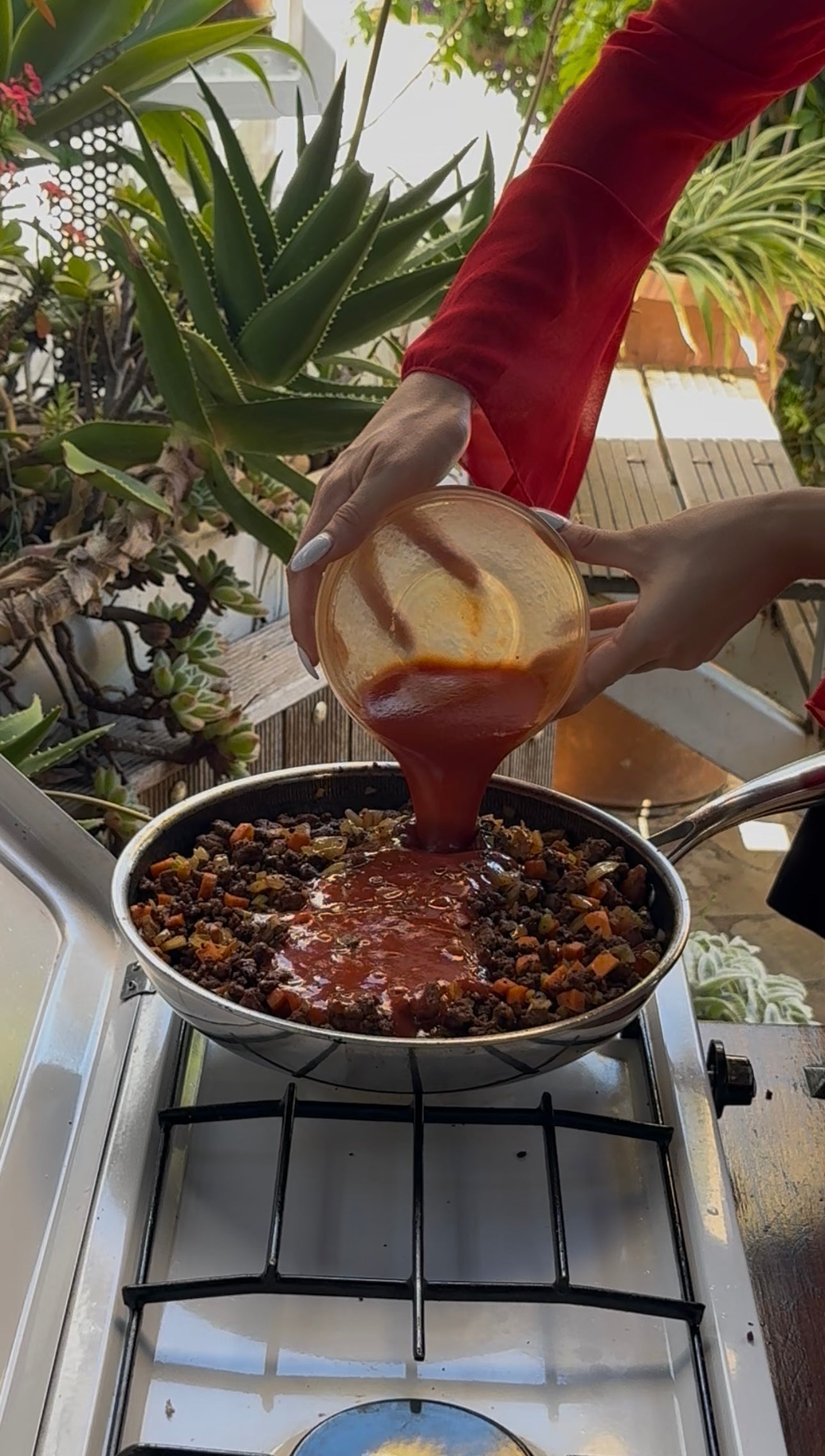 A person pours tomato sauce from a bowl into a skillet of vegetables and ground meat on an outdoor stove, surrounded by lush green plants.