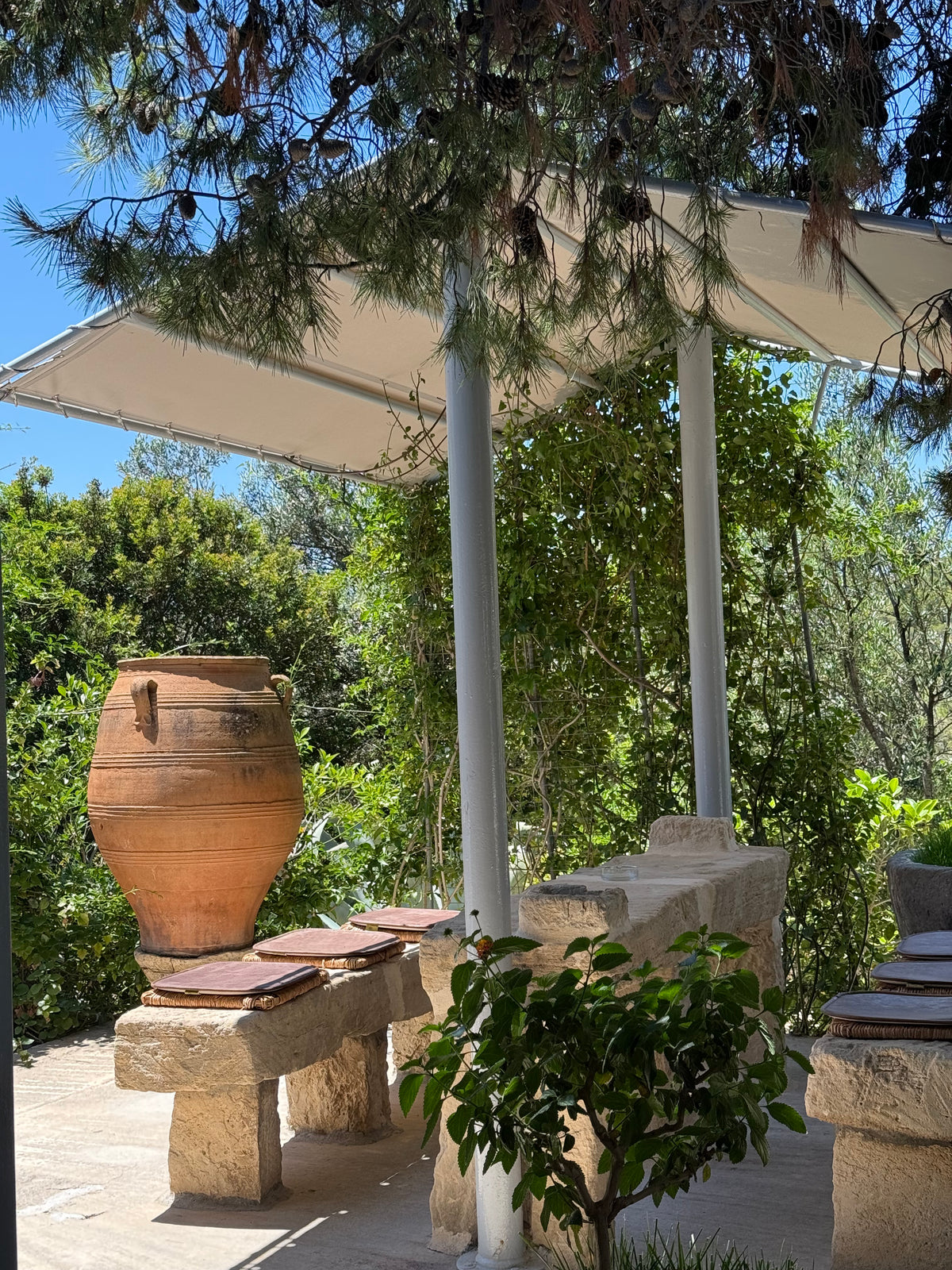 A shaded stone seating area with benches, a large clay pot, lush greenery, and trees surrounding the spot under a white canopy on a sunny day.