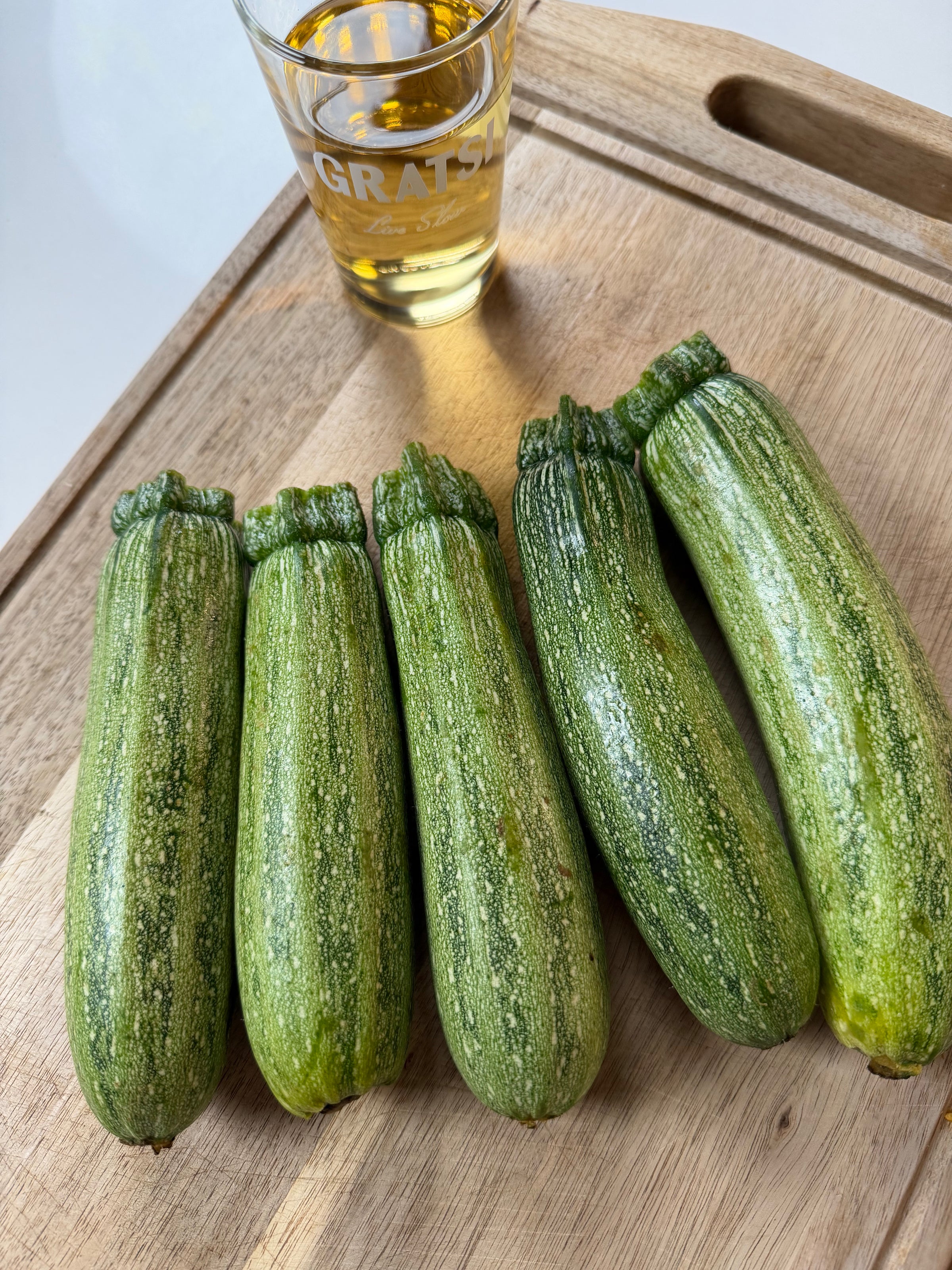 Five zucchinis are lined up on a wooden cutting board next to a glass filled with a golden liquid.