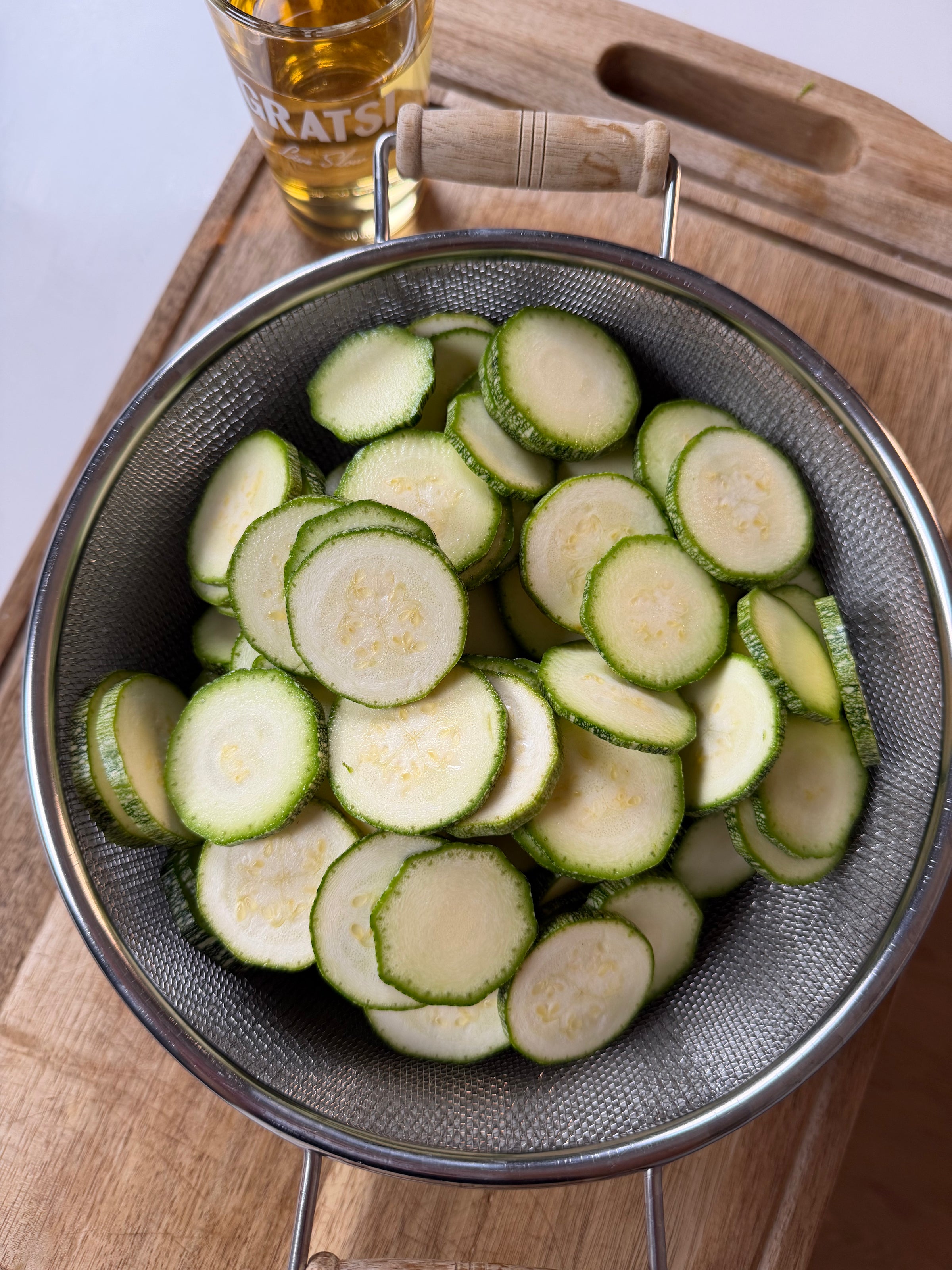 A metal colander filled with sliced zucchini sits on a wooden cutting board. In the background, there is a glass of light-colored liquid, possibly beer.