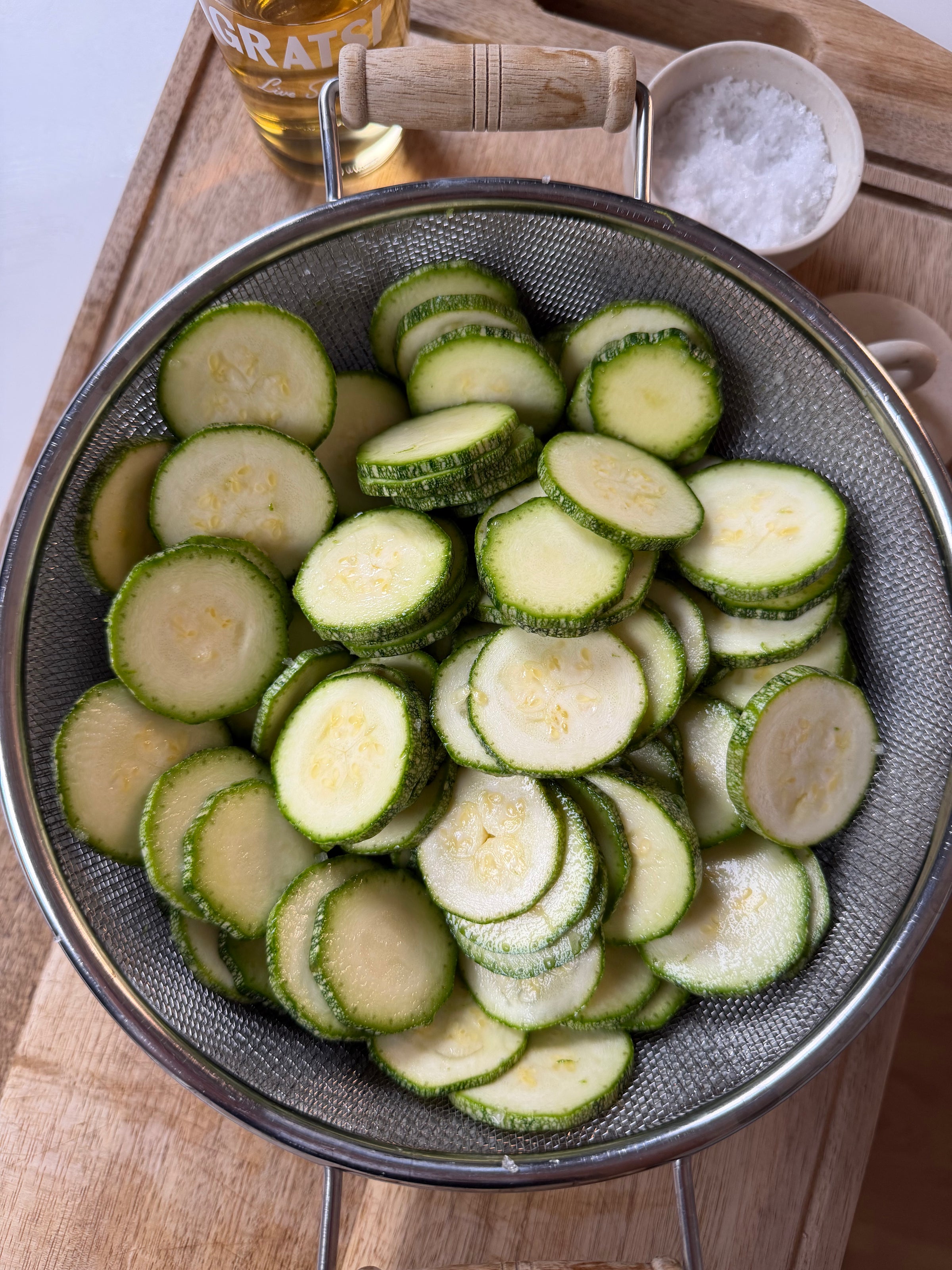 A metal colander filled with sliced zucchini sits on a wooden surface. Nearby, a bowl of coarse salt and a bottle are partially visible.