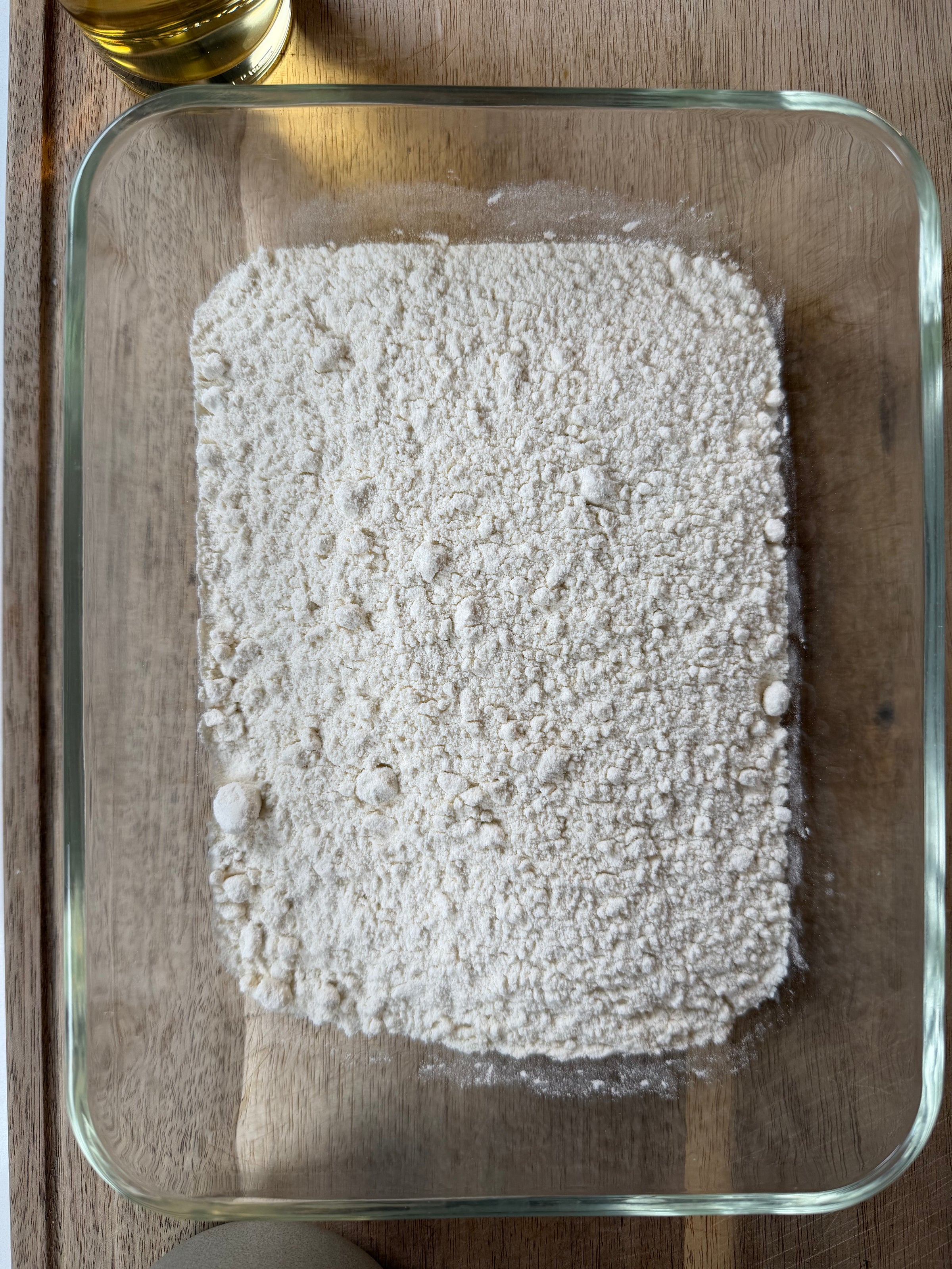 A glass dish filled with white flour sits on a wooden surface. Part of a glass jar with yellow liquid and a round container are partially visible at the top and bottom edges.