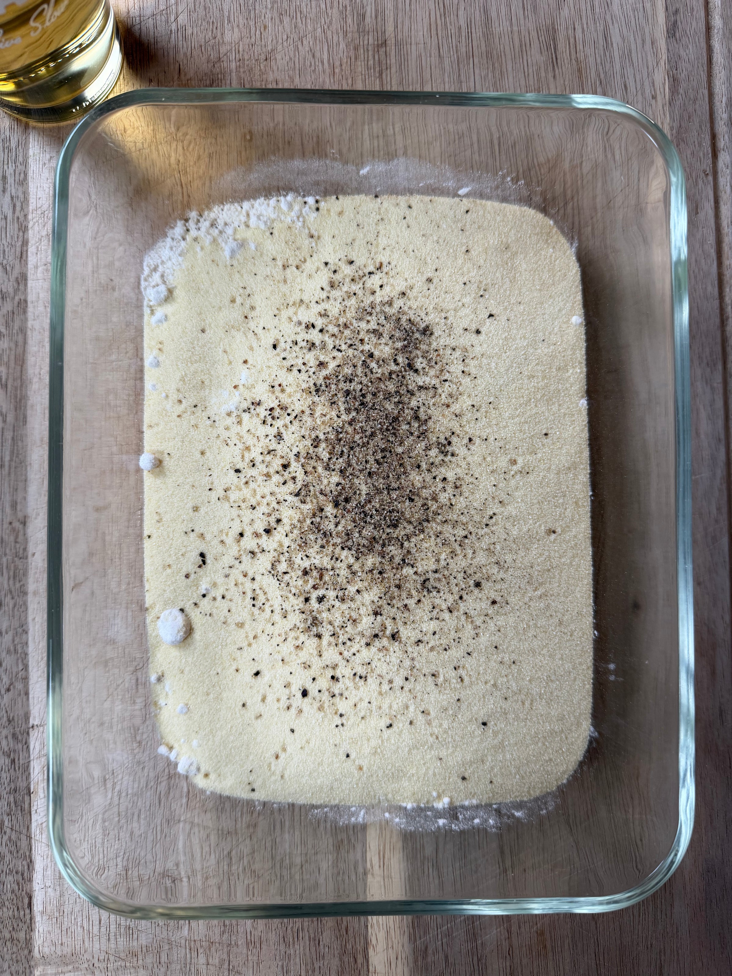 A glass dish filled with a layer of cornmeal topped with ground black pepper, sitting on a wooden surface. A small glass of light yellow liquid is partially visible in the top left corner.