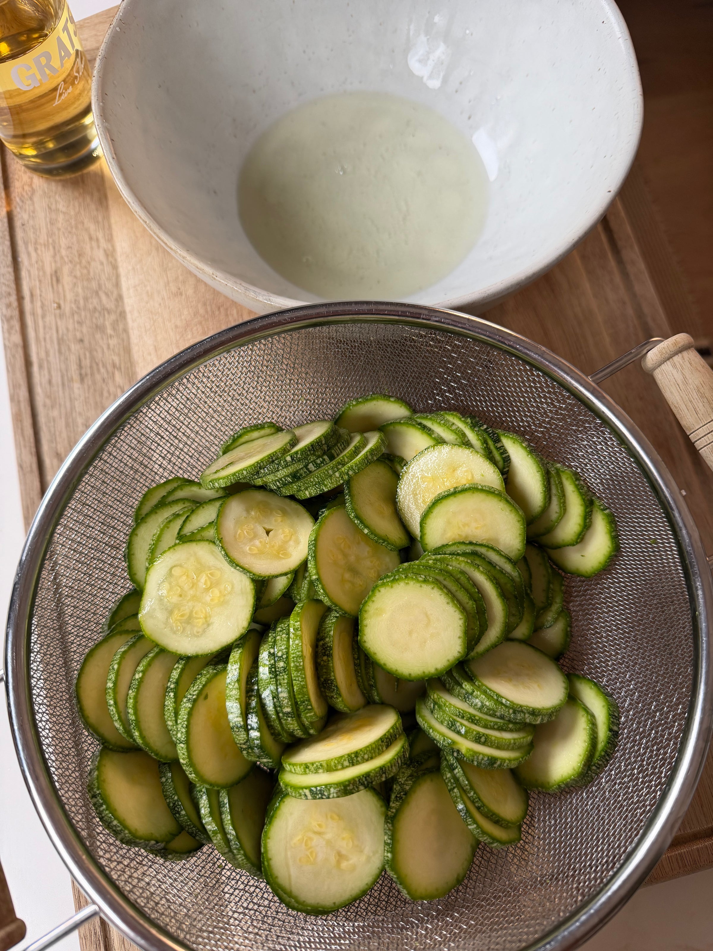 A metal strainer filled with sliced zucchini sits on a wooden surface. Behind it is a white mixing bowl containing a small amount of liquid, with a bottle of olive oil partially visible in the corner.