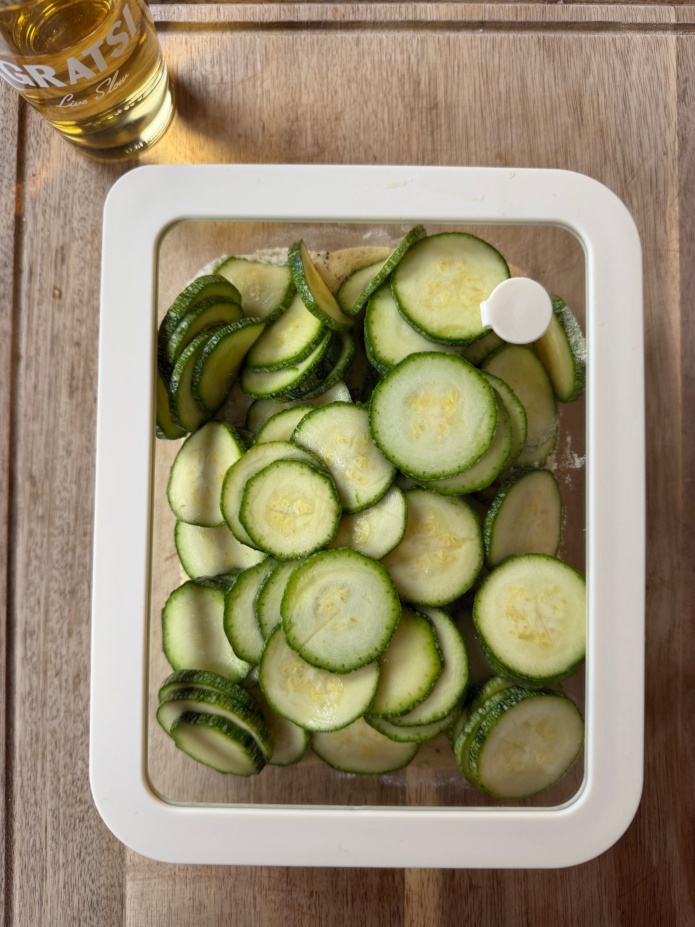 A container filled with thinly sliced cucumbers sits on a wooden surface. In the top left corner, there is a glass of light-colored beverage partly visible with GRATSI written on it.