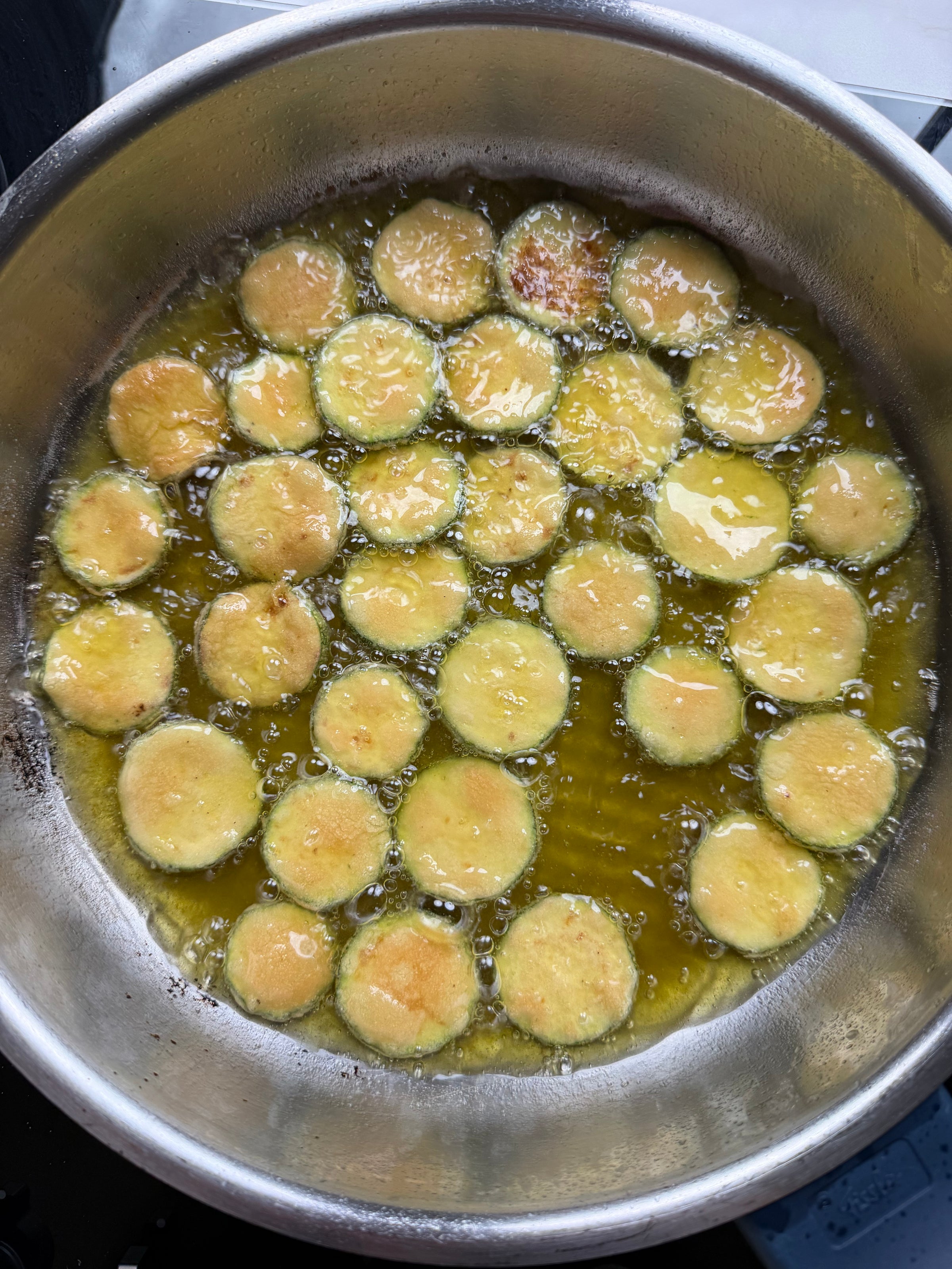 Sliced zucchini rounds frying in hot oil inside a metal pan, with bubbles forming around the vegetables as they cook to a golden brown.