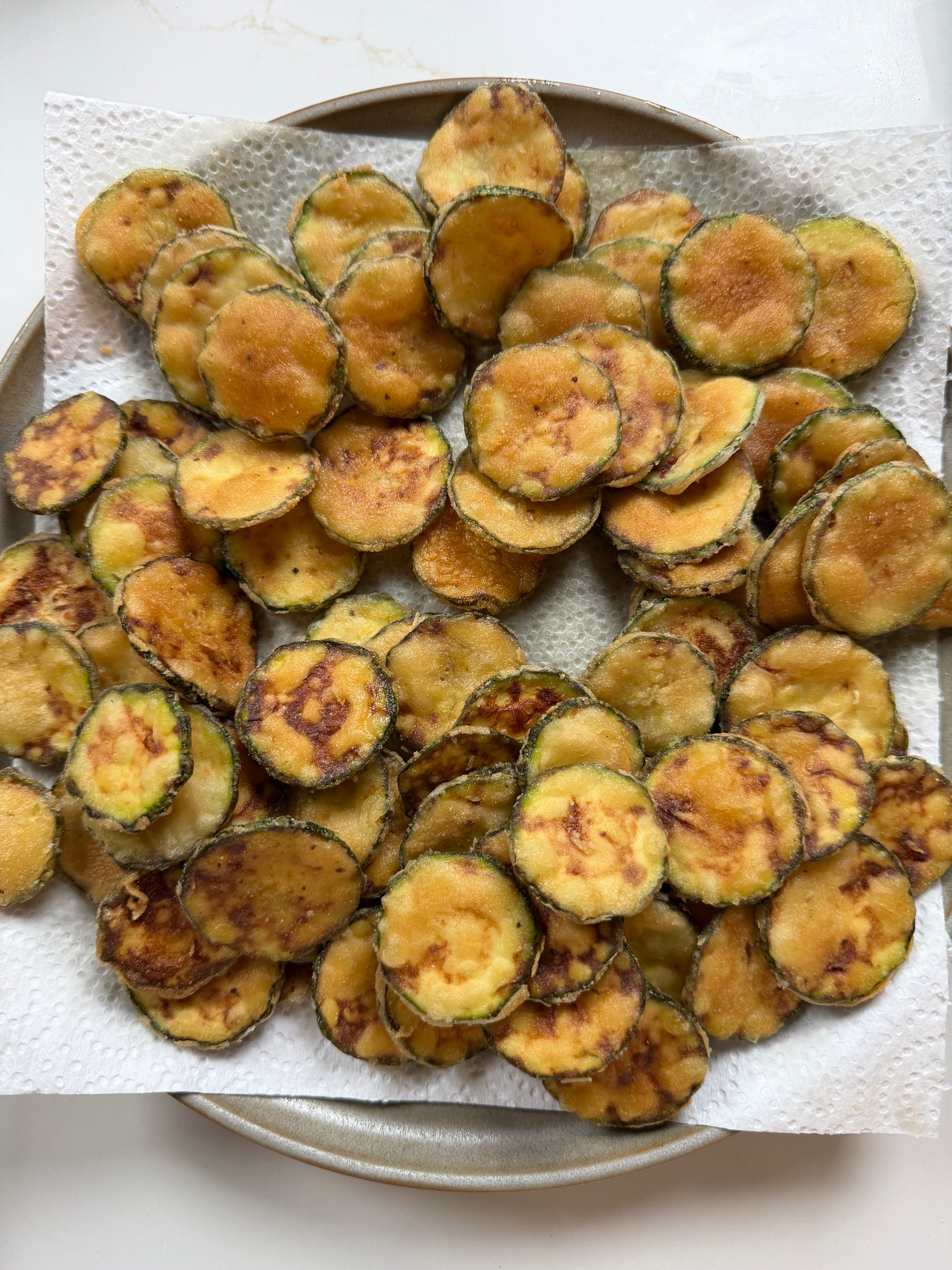 A plate filled with thin, round slices of fried zucchini resting on a paper towel to absorb excess oil. The zucchini slices are golden brown and arranged in a slightly overlapping pattern.