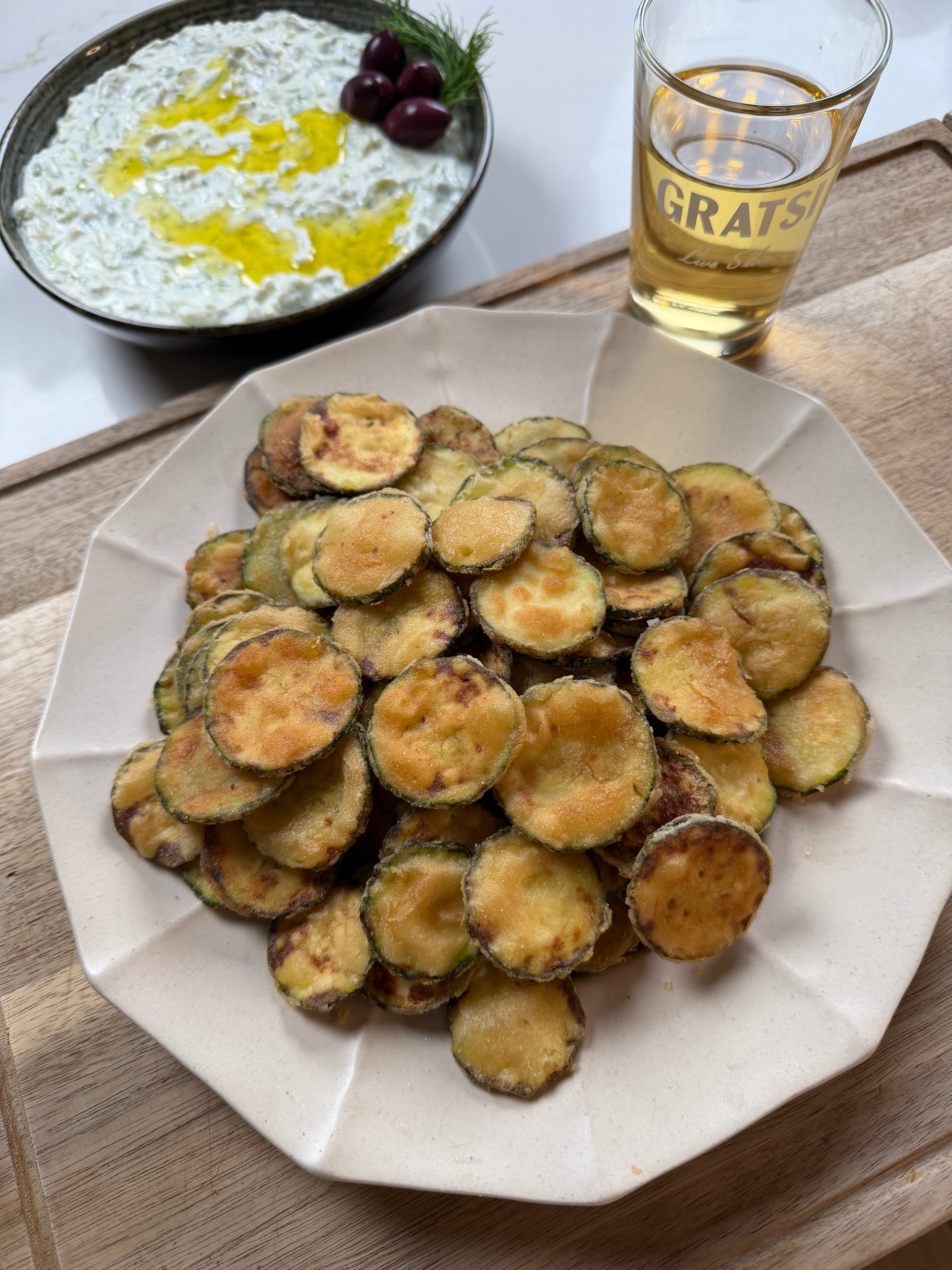 A plate of golden, crispy fried zucchini slices sits in the foreground. Behind it is a bowl of creamy dip garnished with olives and herbs, and a glass of light-colored beverage on a wooden surface.