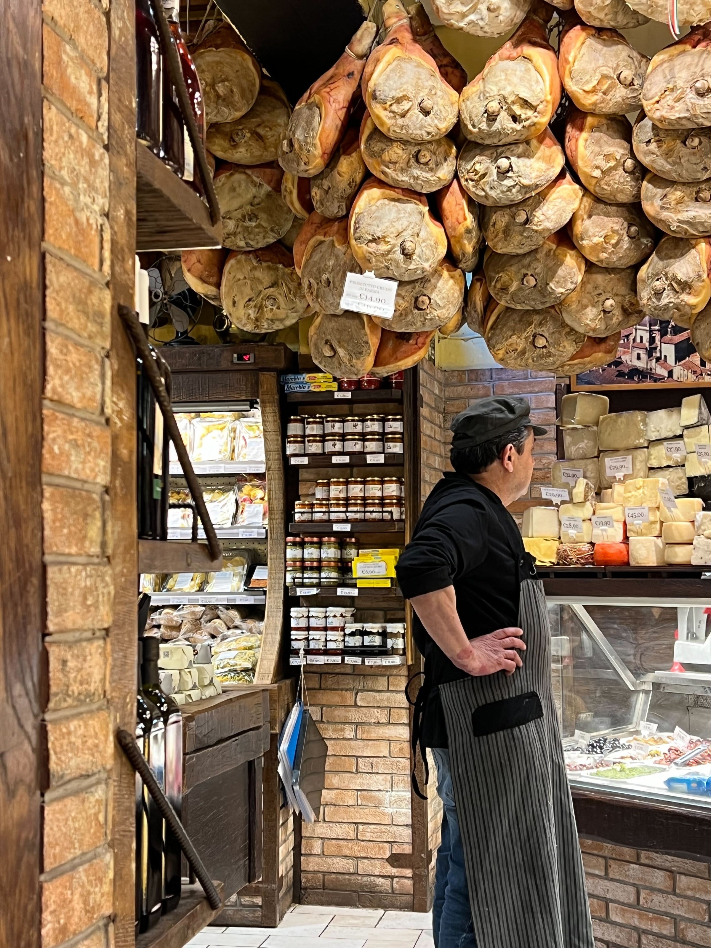 A shop worker in a striped apron stands beside a meat and cheese counter in a rustic deli, with cured hams hanging from the ceiling and shelves filled with jars and cheeses lining the brick walls.