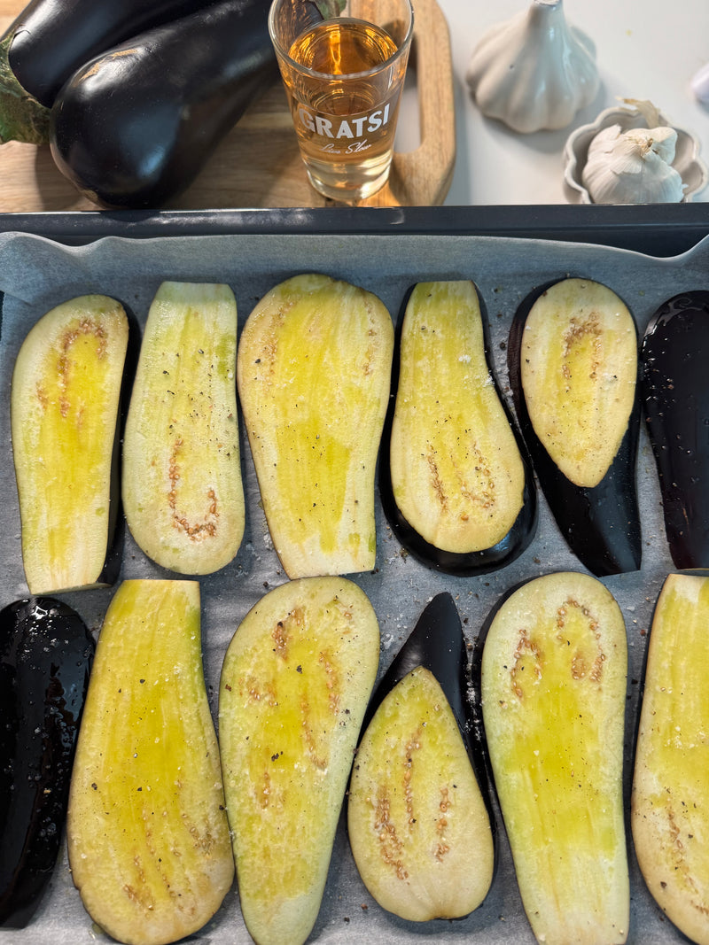 Sliced eggplant drizzled with oil and seasoning on a baking tray, ready to be roasted. In the background are whole eggplants, a glass of amber liquid, a bulb of garlic, and a wooden cutting board.